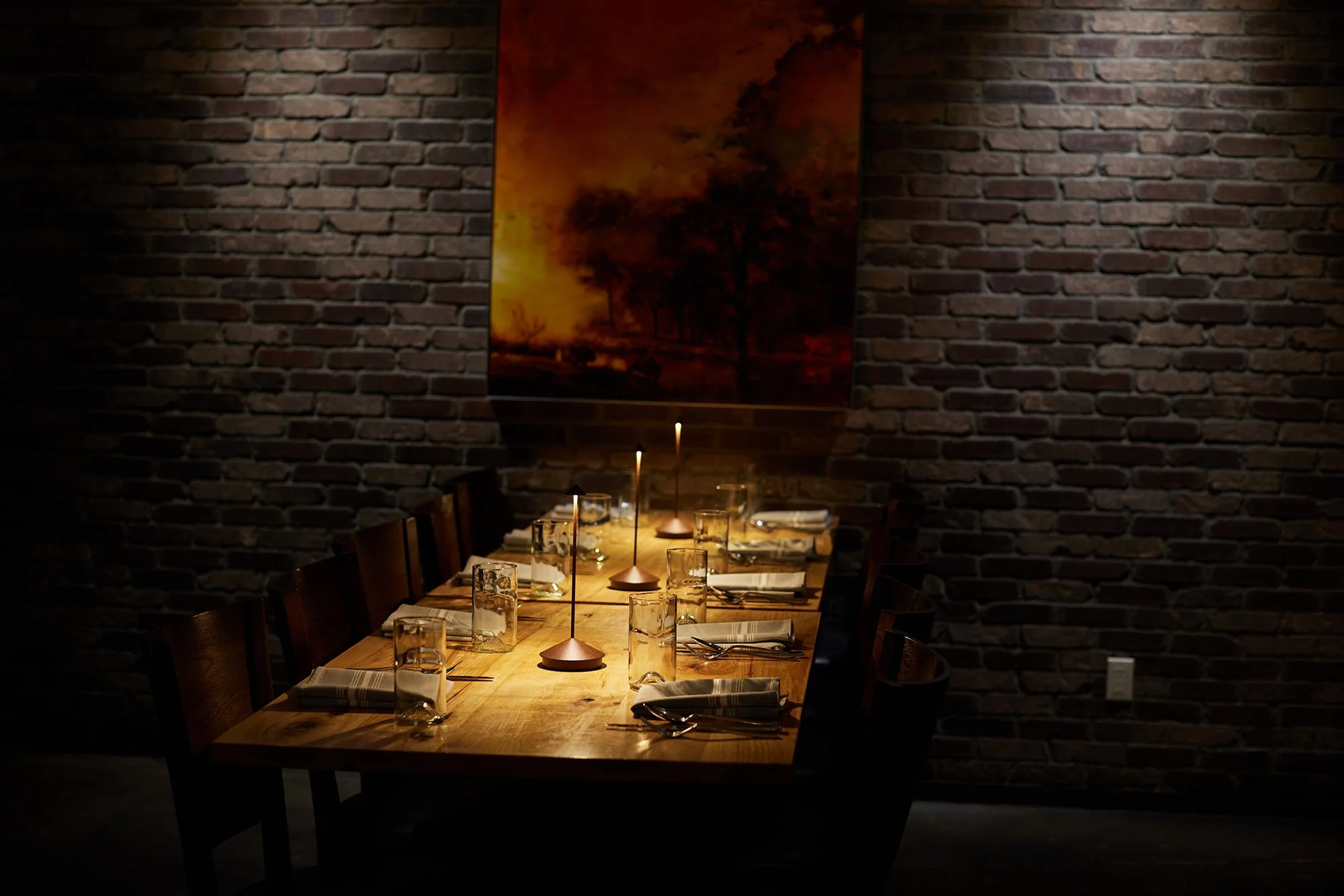 A cozy dining room with a long wooden table set with glasses and napkins, lit by small table lamps, against a brick wall with a large abstract painting.