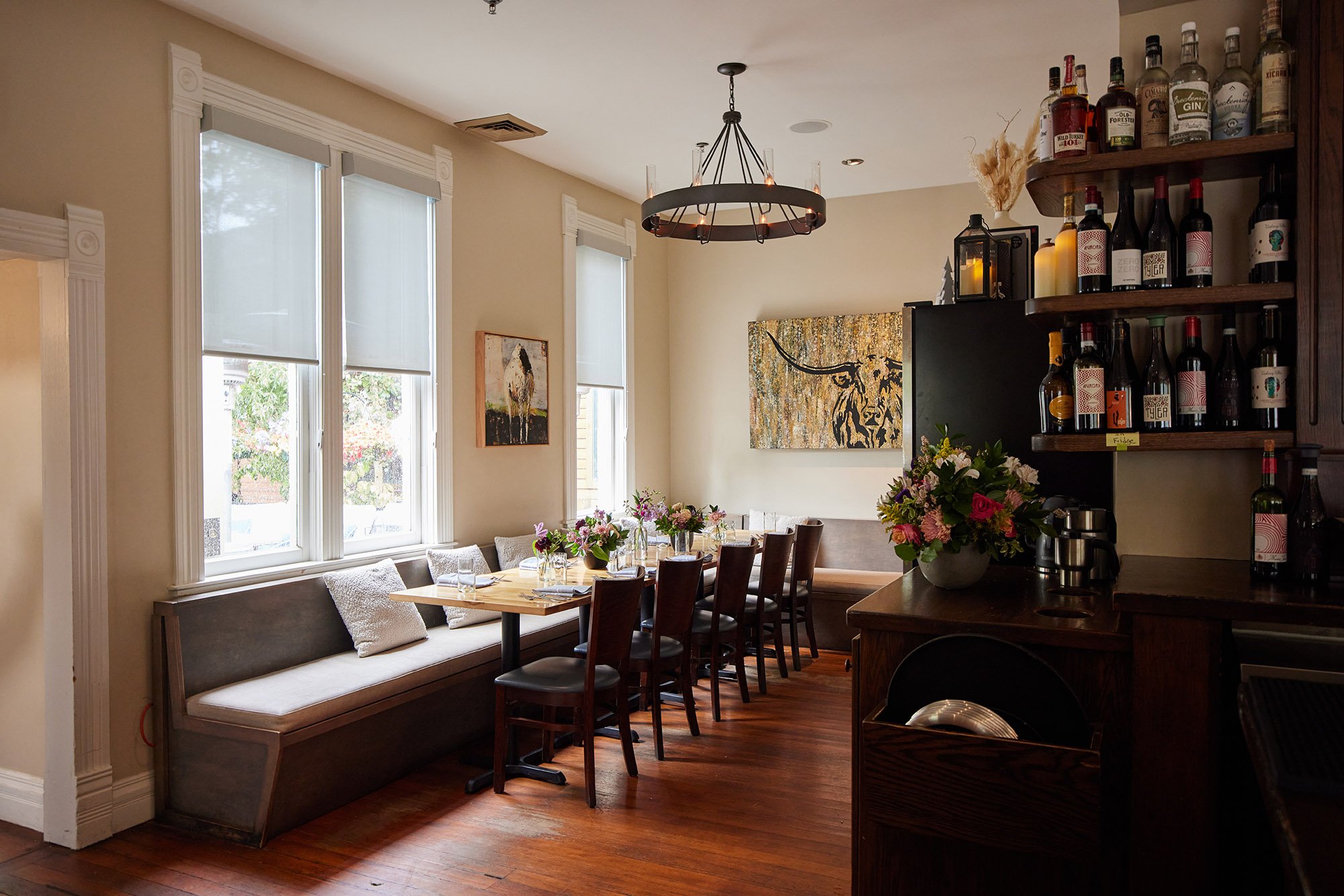 Bright dining room with three large windows with white trim, beige walls, and decorative crown molding. A long built-in bench with pillows lines the windows, and a wooden dining table with chairs and floral centerpieces is in the center. Artworks ado