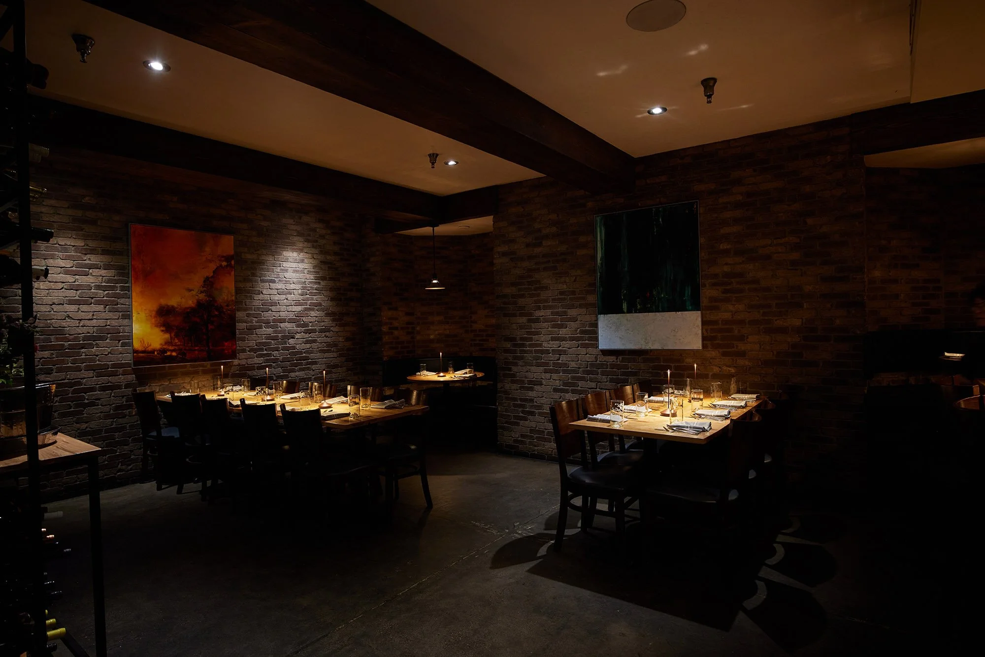 A dimly lit restaurant with brick walls, two paintings on the walls, wooden tables set with glasses, plates, and candles, and chairs around the tables.