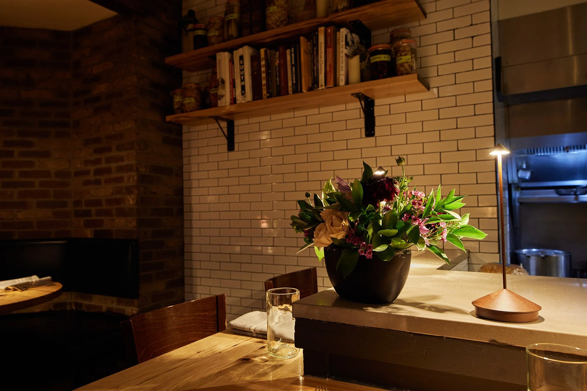 A dining area with a dark wooden table, a glass of water, a white napkin, and a flower arrangement with pink and purple flowers in a black vase on a countertop. Wooden shelves with books and jars are mounted on a white brick wall in the background. A