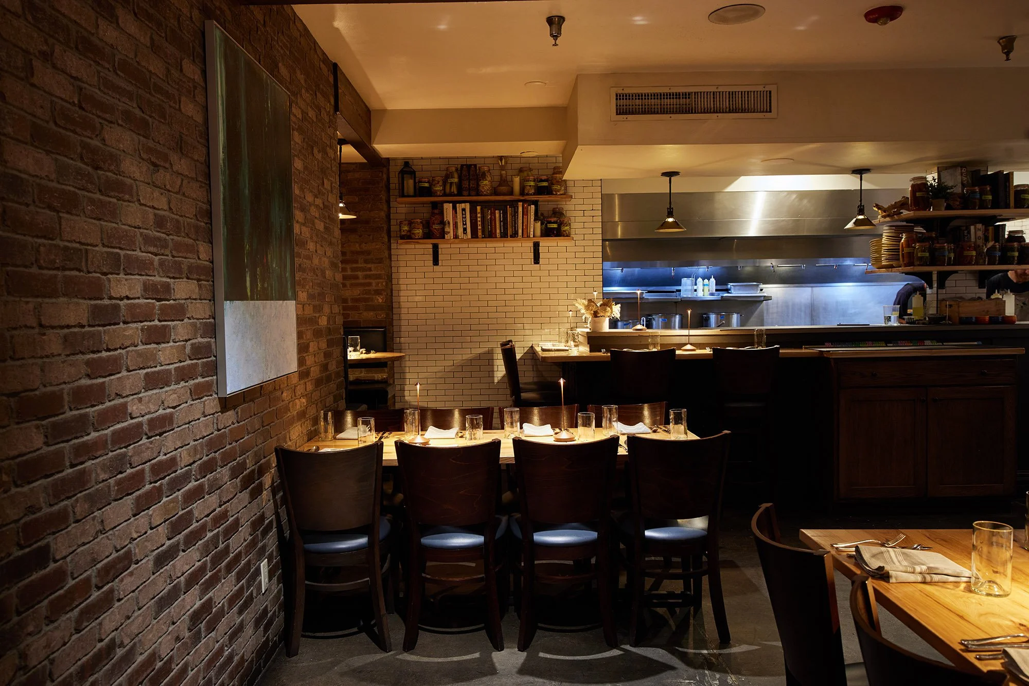 A cozy restaurant interior with exposed brick walls, wooden tables, and dark wooden chairs. The table in the foreground is set with glasses and napkins. In the background, there is a kitchen area with stainless steel appliances and shelves stocked wi