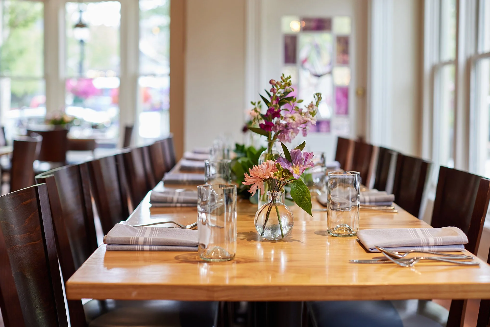 A dining table set with a centerpiece of pink and purple flowers in glass vases, glasses, napkins, and silverware, in a bright room with large windows and wooden chairs.