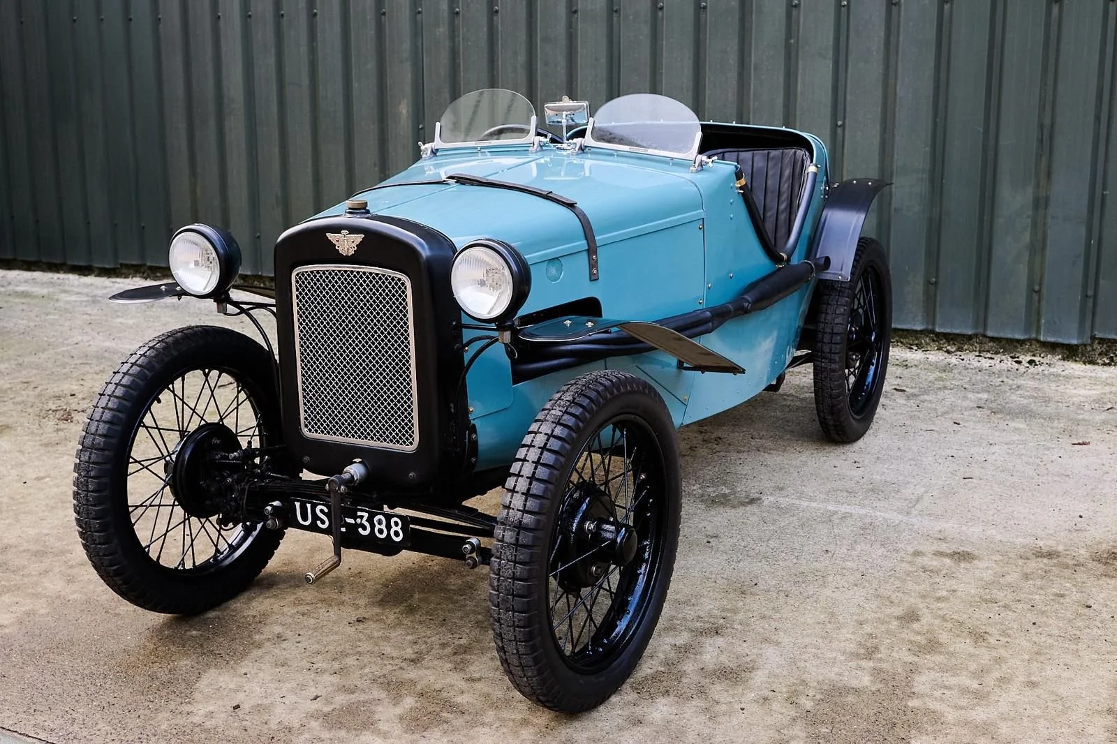 A vintage blue and black open-top automobile with a single row of seating, large headlights, and black wire wheels, parked on a dirt surface in front of a corrugated metal building.