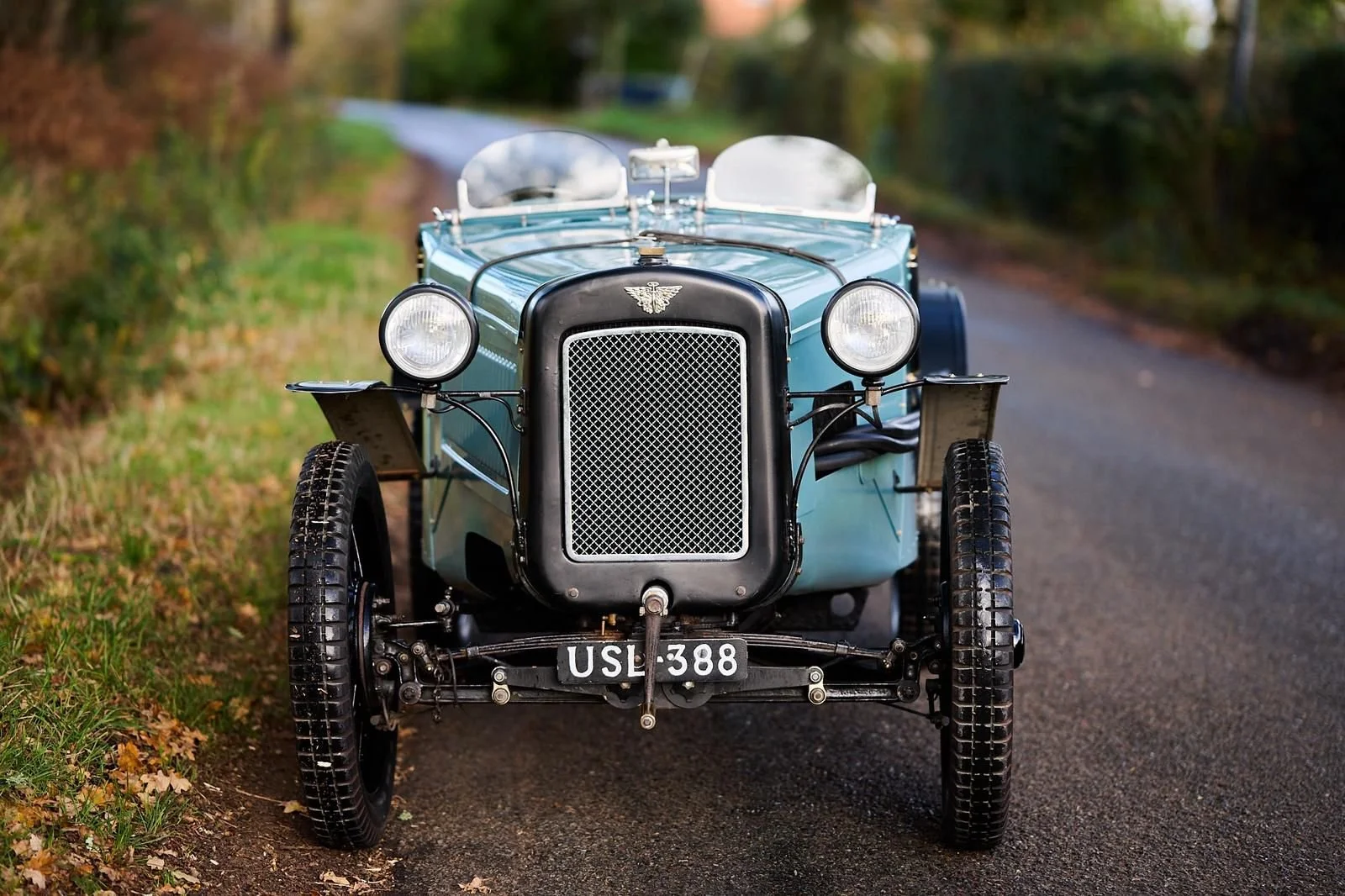 Front view of a vintage light blue open-top car with round headlights, a black radiator grille, and a license plate reading 'USE 388', parked on a narrow country road.
