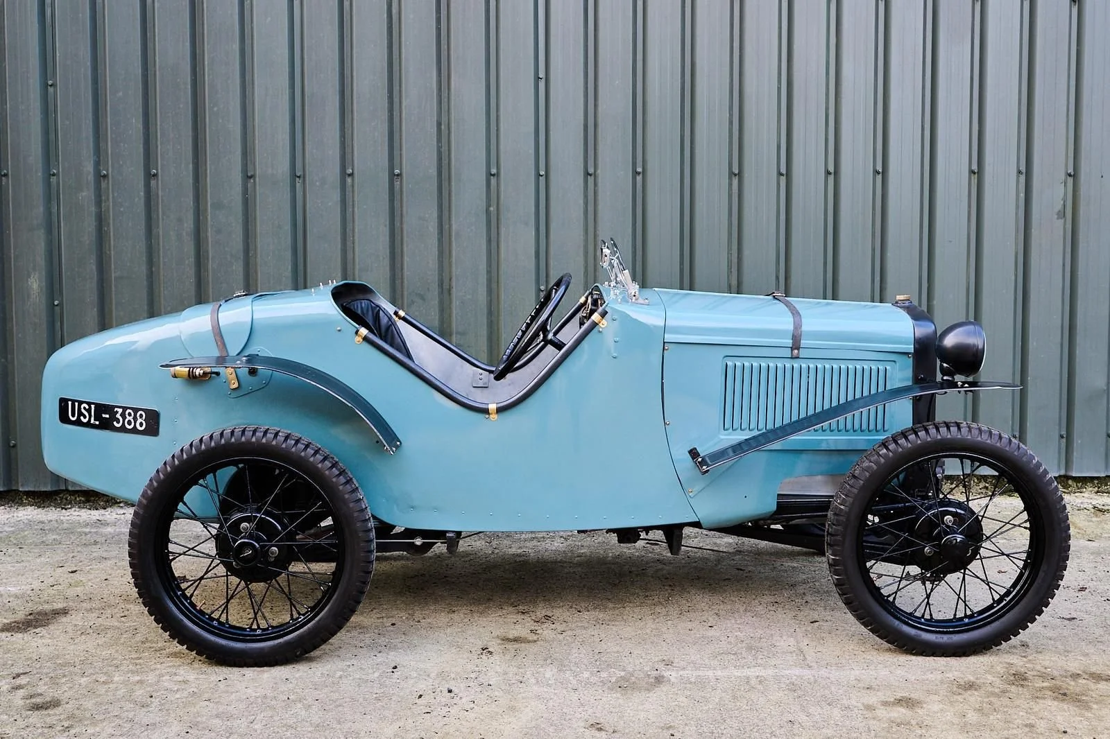 A vintage, open-top race car painted in light blue with black wire-spoked wheels, parked on a dirt ground in front of a metal wall.