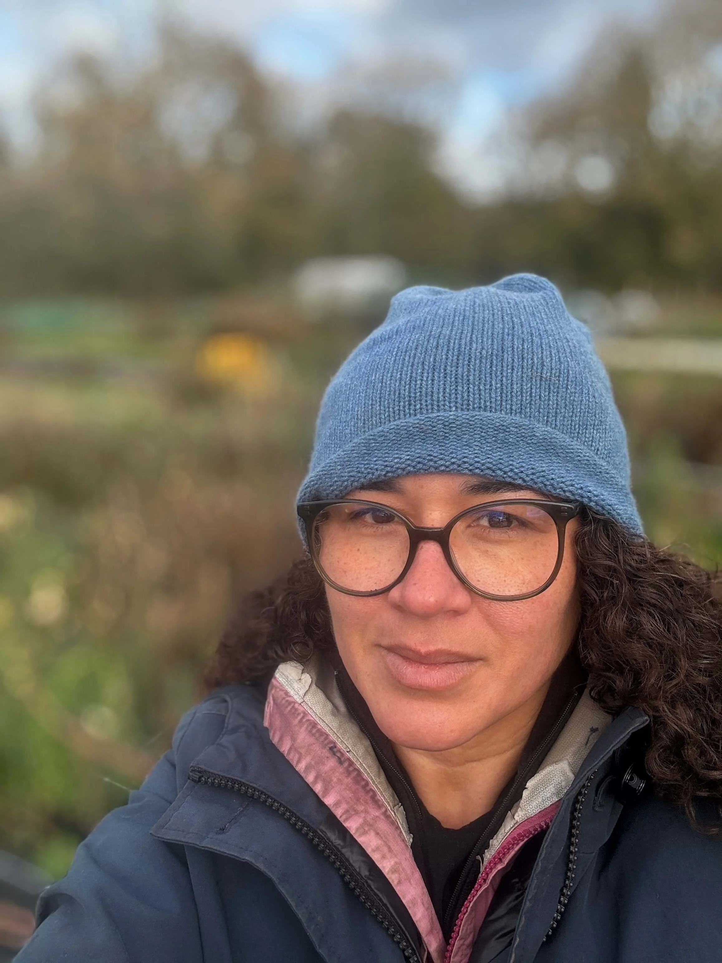 A woman outdoors wearing a blue knit beanie, glasses, and a jacket. She has curly hair and appears to be in a park or natural setting with blurred trees and foliage in the background.