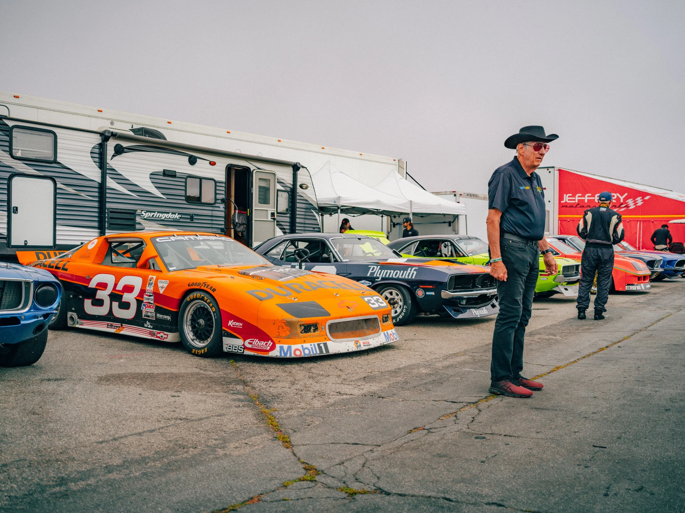 Race cars parked in a pit area alongside trailers and tents with people walking around, in what appears to be a racing event setup.