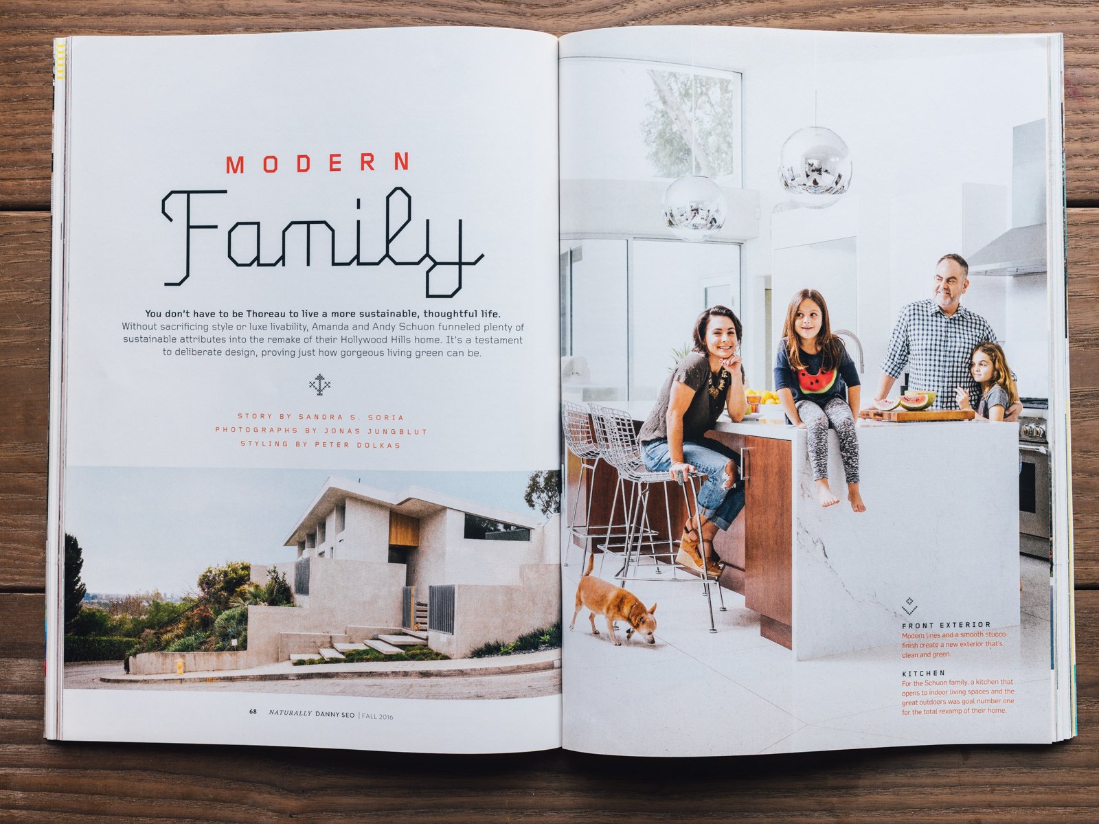 An open magazine spread showing a modern family in a bright, white kitchen with high ceilings and hanging pendant lights. The family, including two women, a man, and two children, are gathered around a kitchen island, smiling and engaging with each o