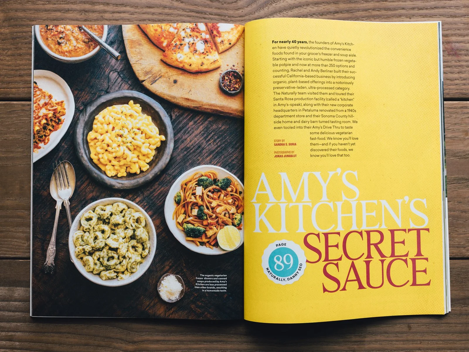 Open magazine with a two-page spread; left page shows a wooden table with various bowls of pasta, salad, and chips, along with pizza on a wooden cutting board; right page has yellow background with black and red text about Amy's Kitchen's secret sauc