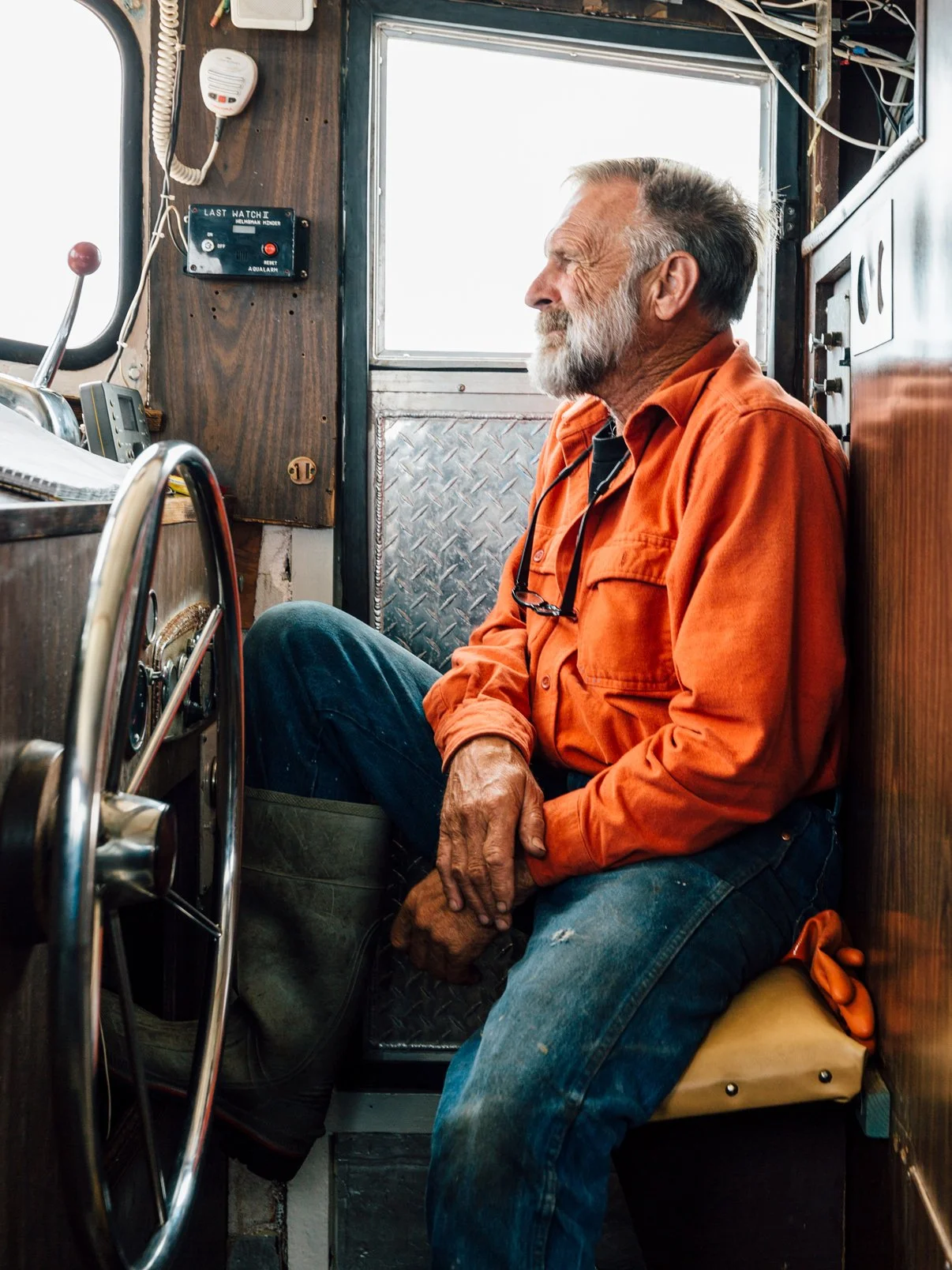 An elderly man with gray hair and a beard, wearing an orange shirt and jeans, sitting in the cabin of a boat or ship, resting with his arms crossed and eyes closed, surrounded by boat controls and equipment.
