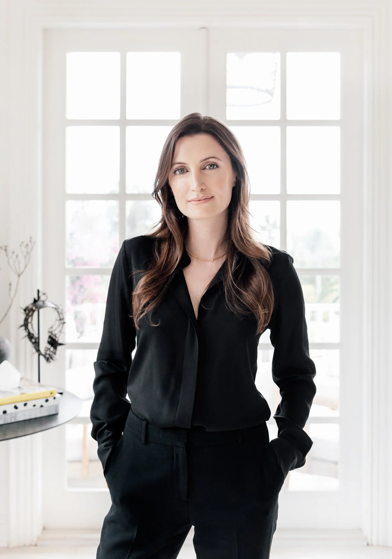 Woman with brown hair wearing a black suit standing in a bright room with large windows