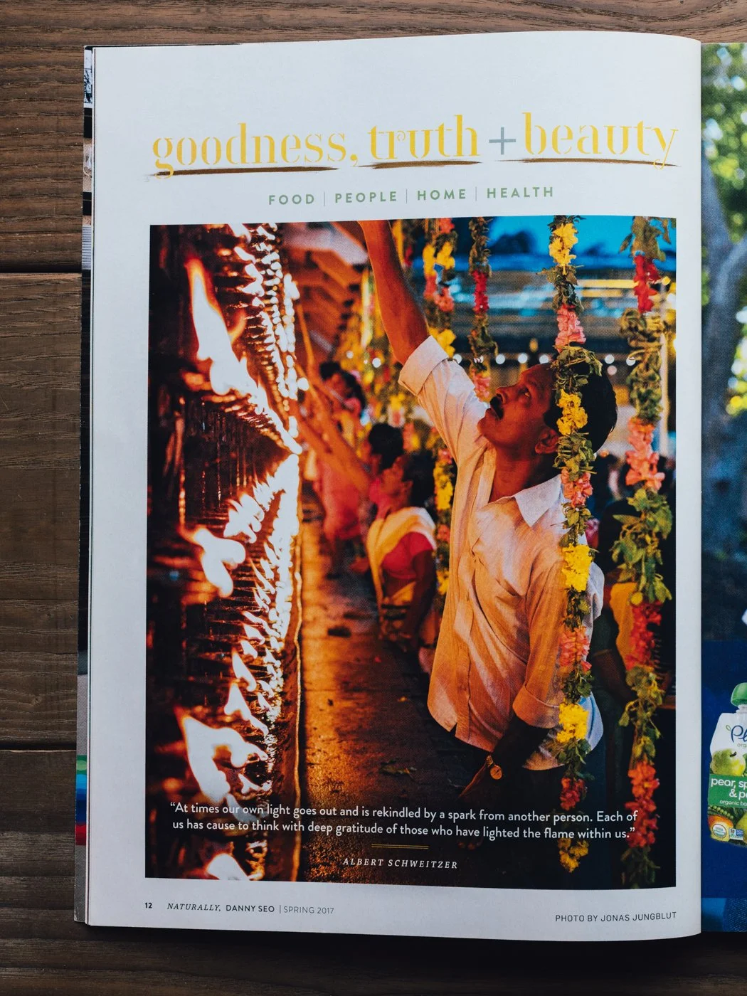 Magazine page showing a man lighting a flame at a festival, decorated with colorful flowers hanging from strings.