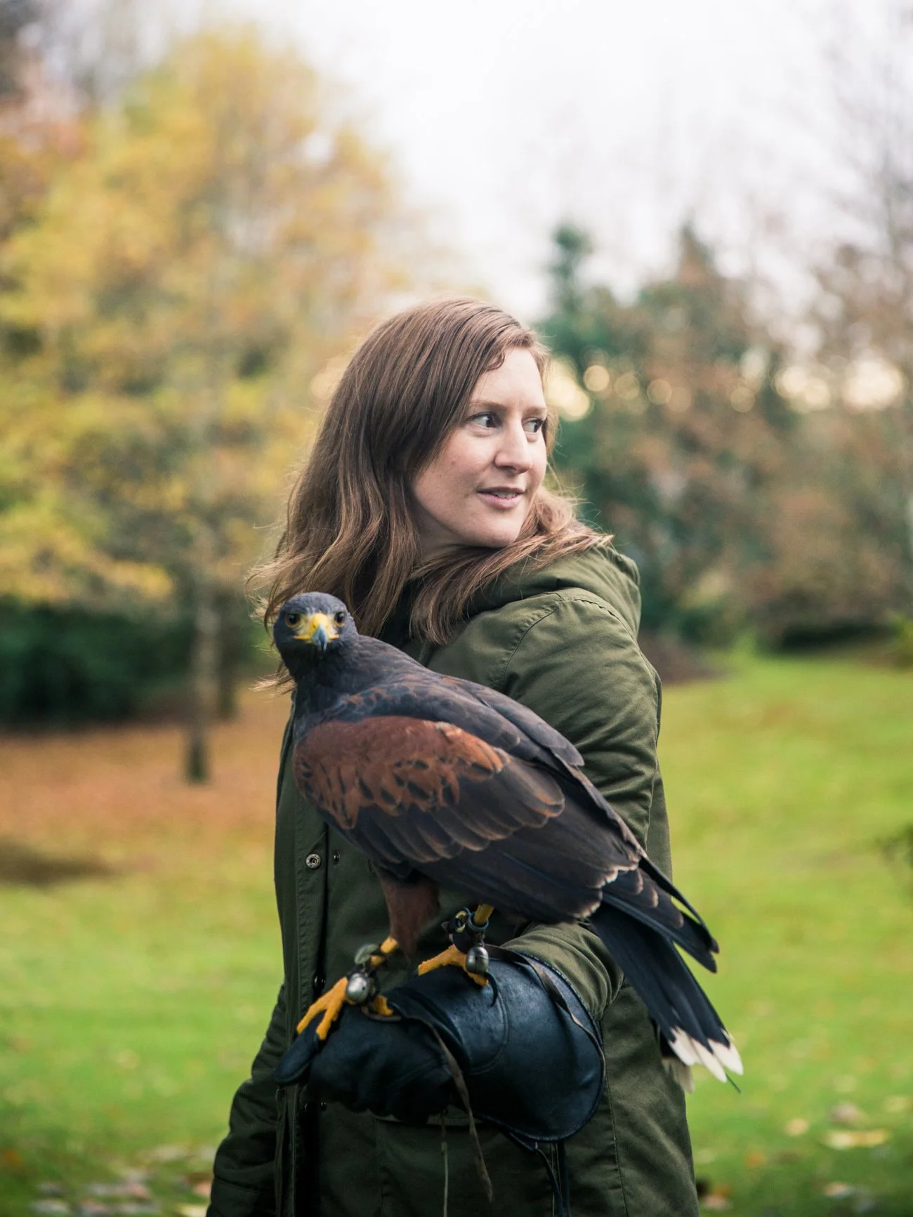 A woman with brown hair wearing a green jacket holding a Harris's hawk outdoors with trees in the background.