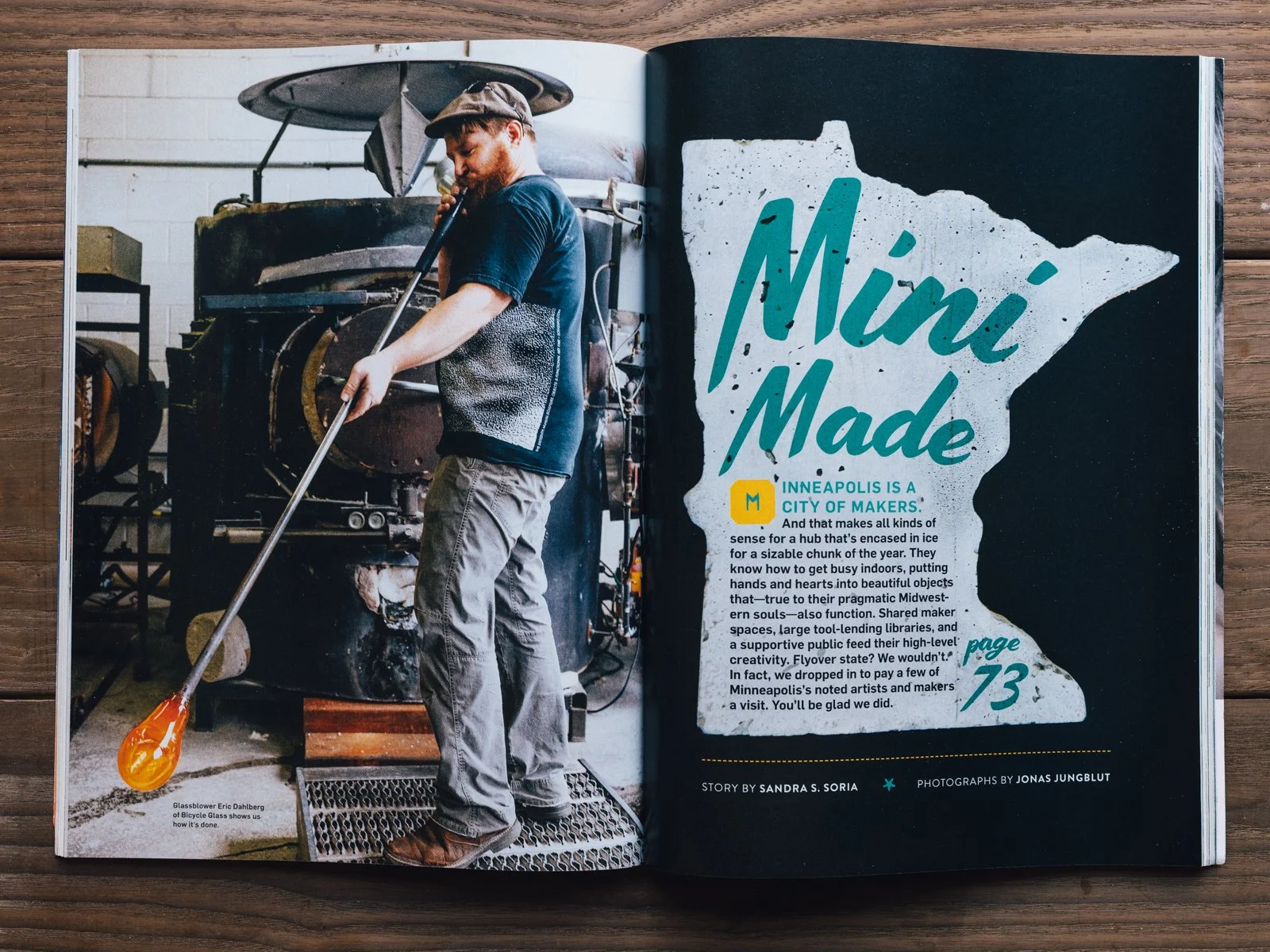 A man working in a workshop using a blowtorch on a glass object, with industrial equipment in the background and a two-page magazine spread about Minneapolis made crafts and makers.