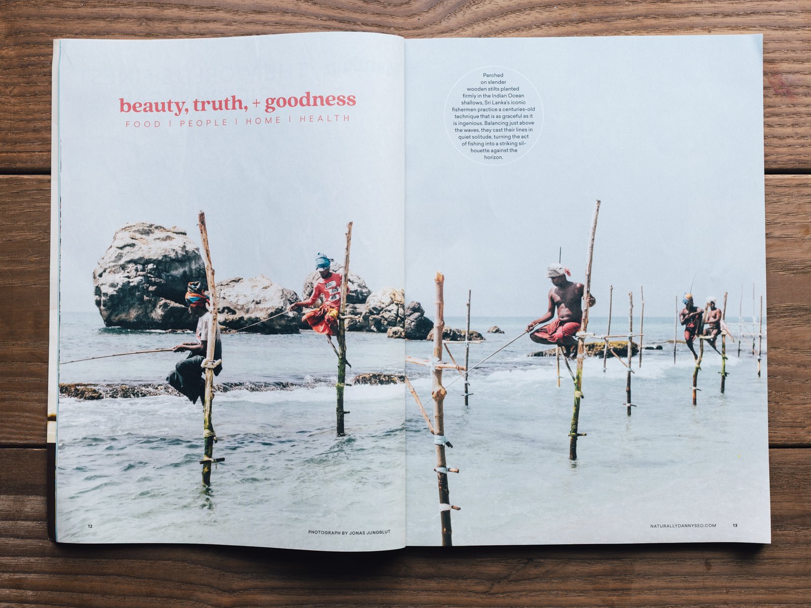 Photograph of people balancing on stilts in the ocean near rocks, with a blue sky overhead.