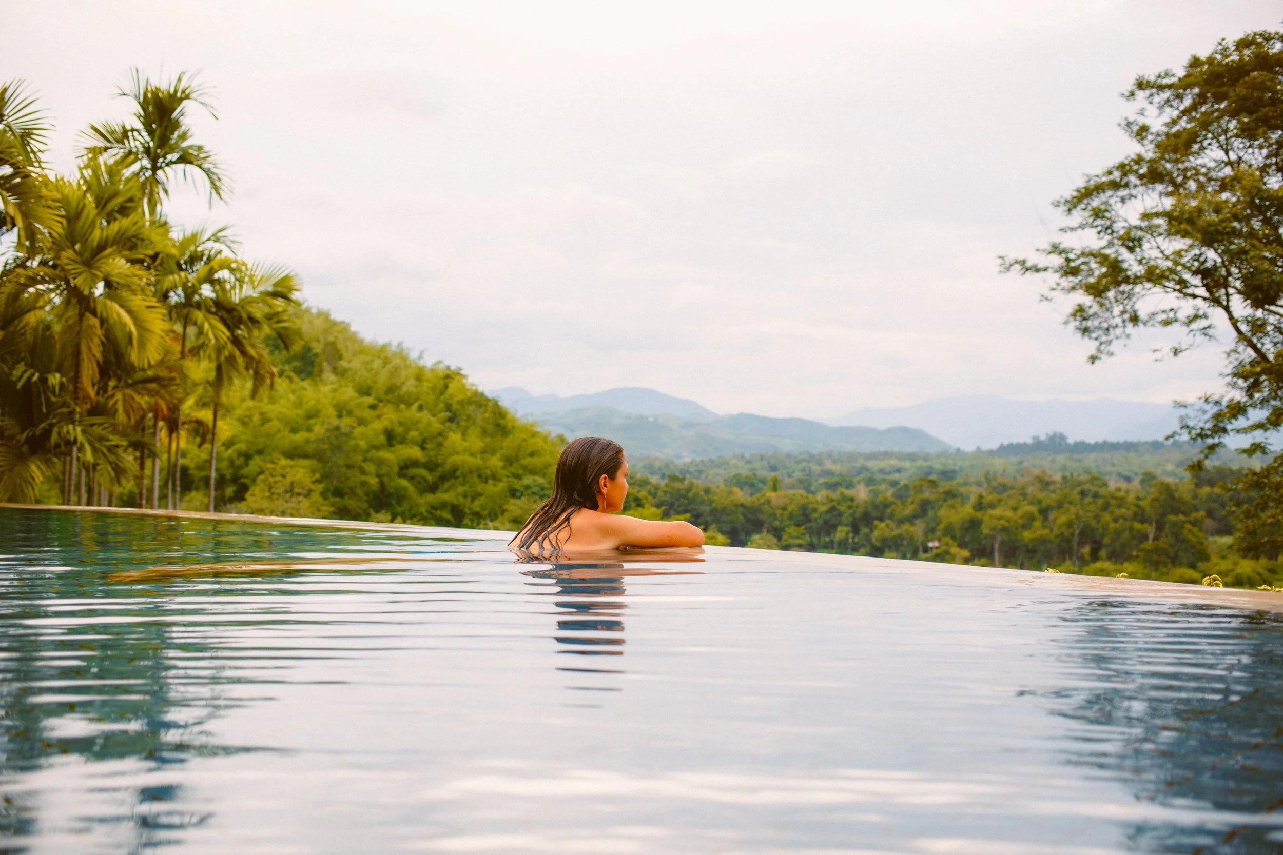 A woman with dark hair is relaxing in an infinity pool at the Anantara Golden Triangle Resort, overlooking lush green hills and trees under a cloudy sky.