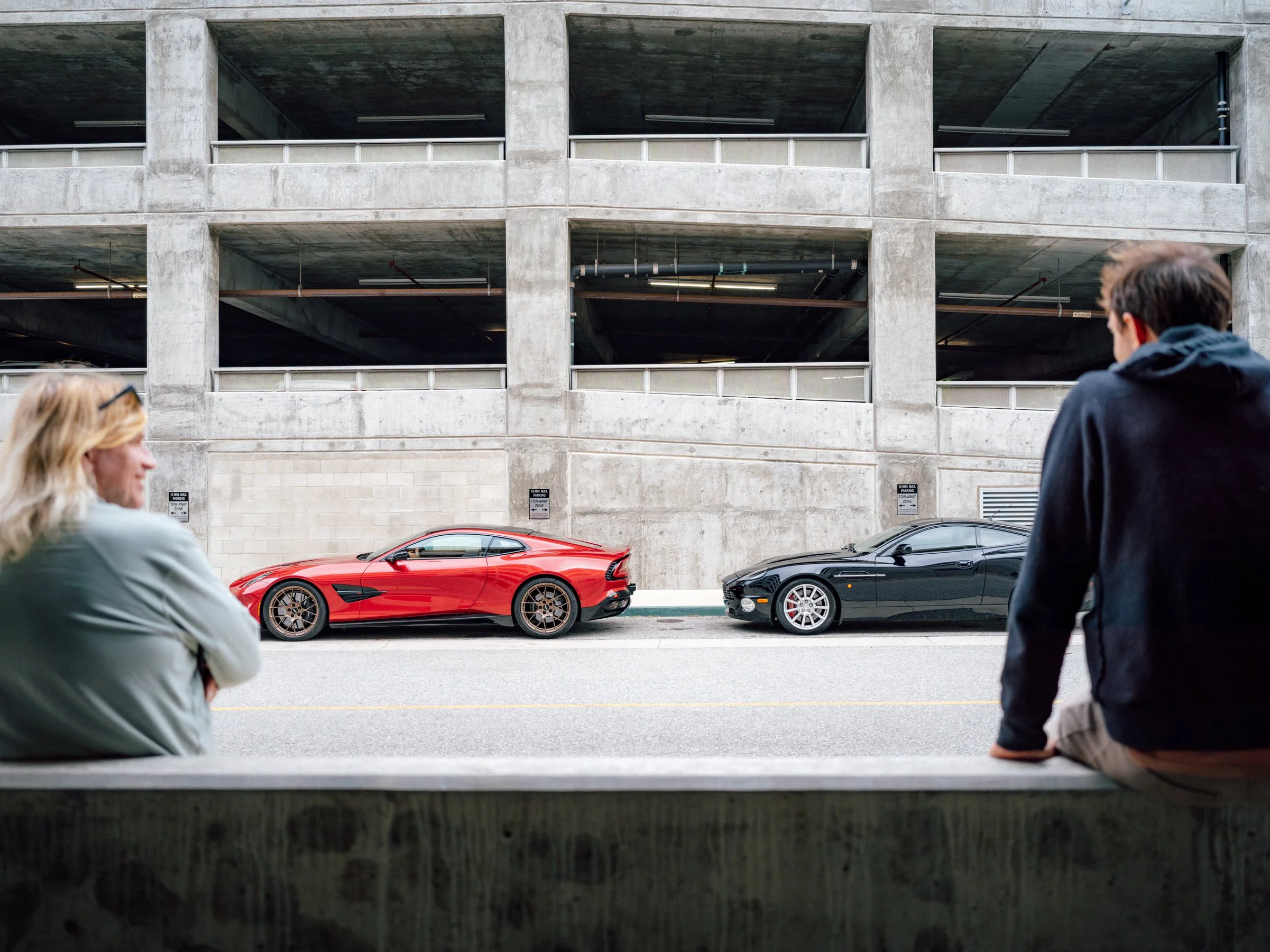 Viewed from under a parking garage, a red sports car and a black sports car are parked in front of a concrete building. Two people are sitting on a ledge in the foreground.