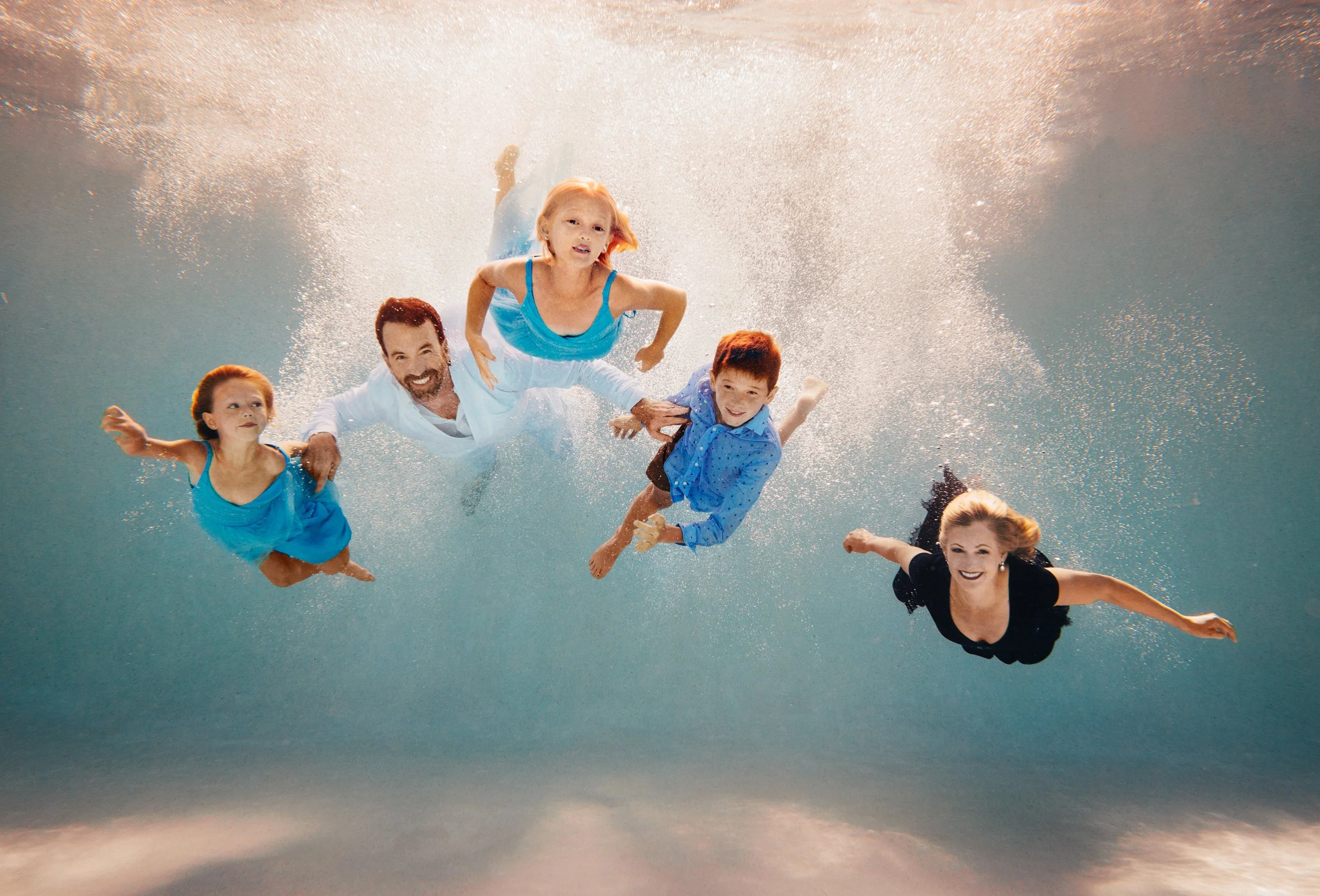 A family of five, including three children and two adults, swimming underwater with bubbles and sunlight filtering through the water.