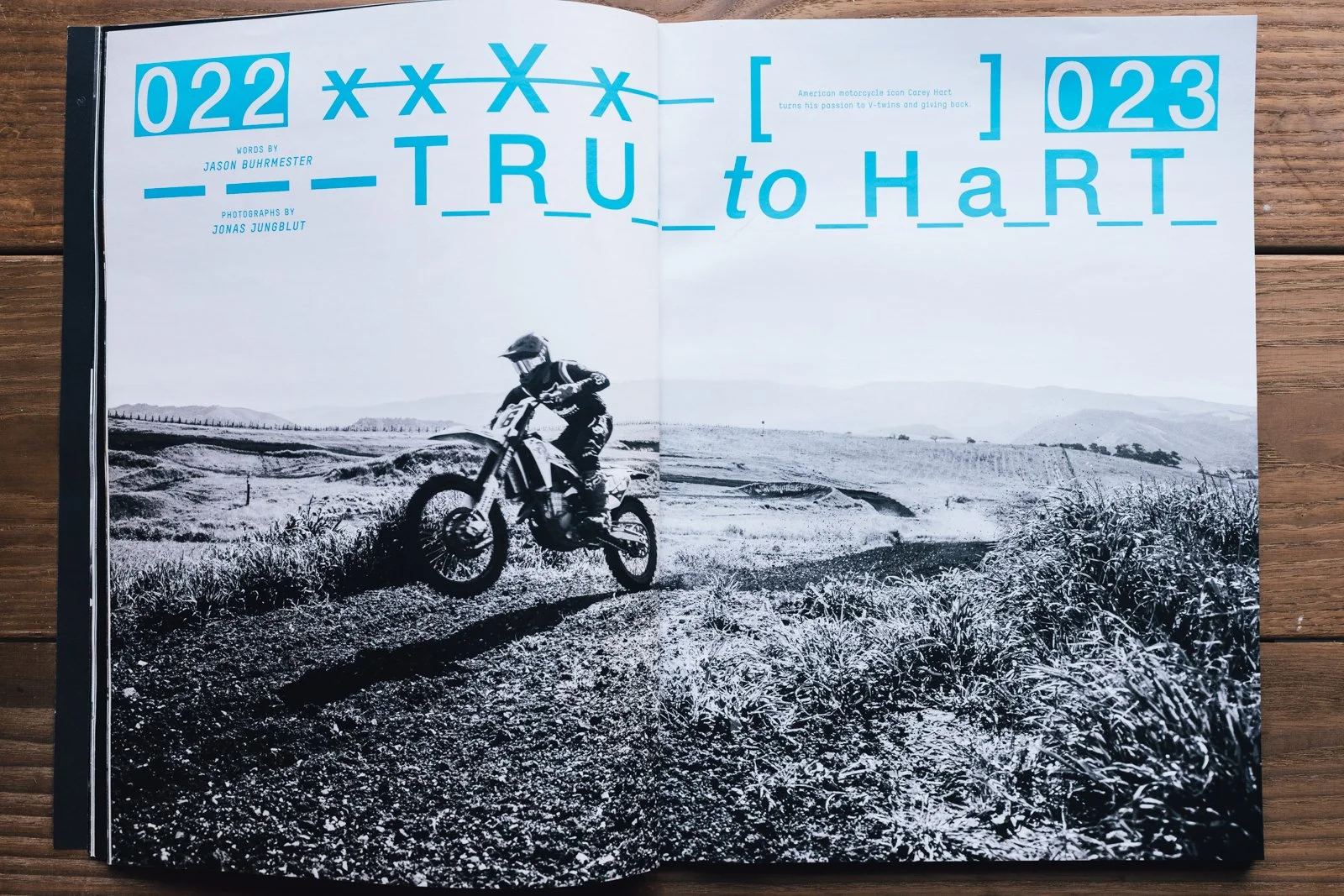 A black-and-white photograph of a person riding a dirt bike on a trail in a rural landscape with rolling hills and some trees in the distance.
