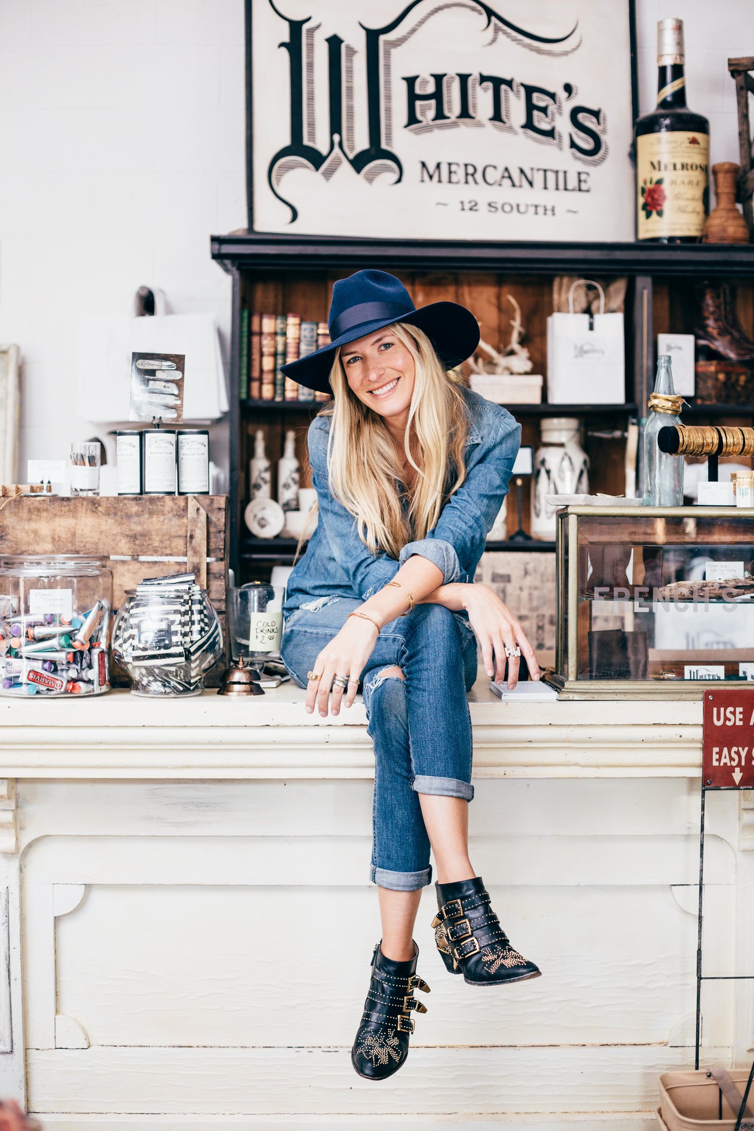 A smiling woman wearing a blue wide-brim hat, denim jacket, and distressed jeans, sitting on a shelf with her left leg crossed over her right, in a vintage shop.