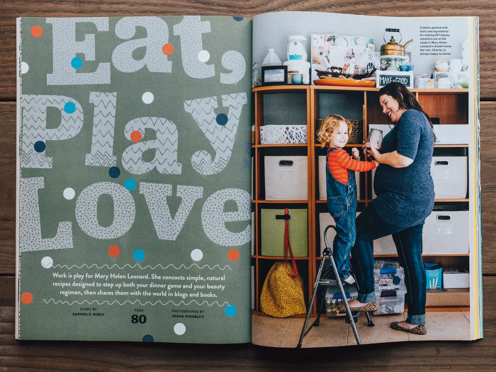 A woman and a young boy interact in front of a wooden storage unit filled with colorful bins and household items. The woman kneels, holding a jar, while the boy stands on a small step stool, smiling and reaching out to her. The setting appears to be 