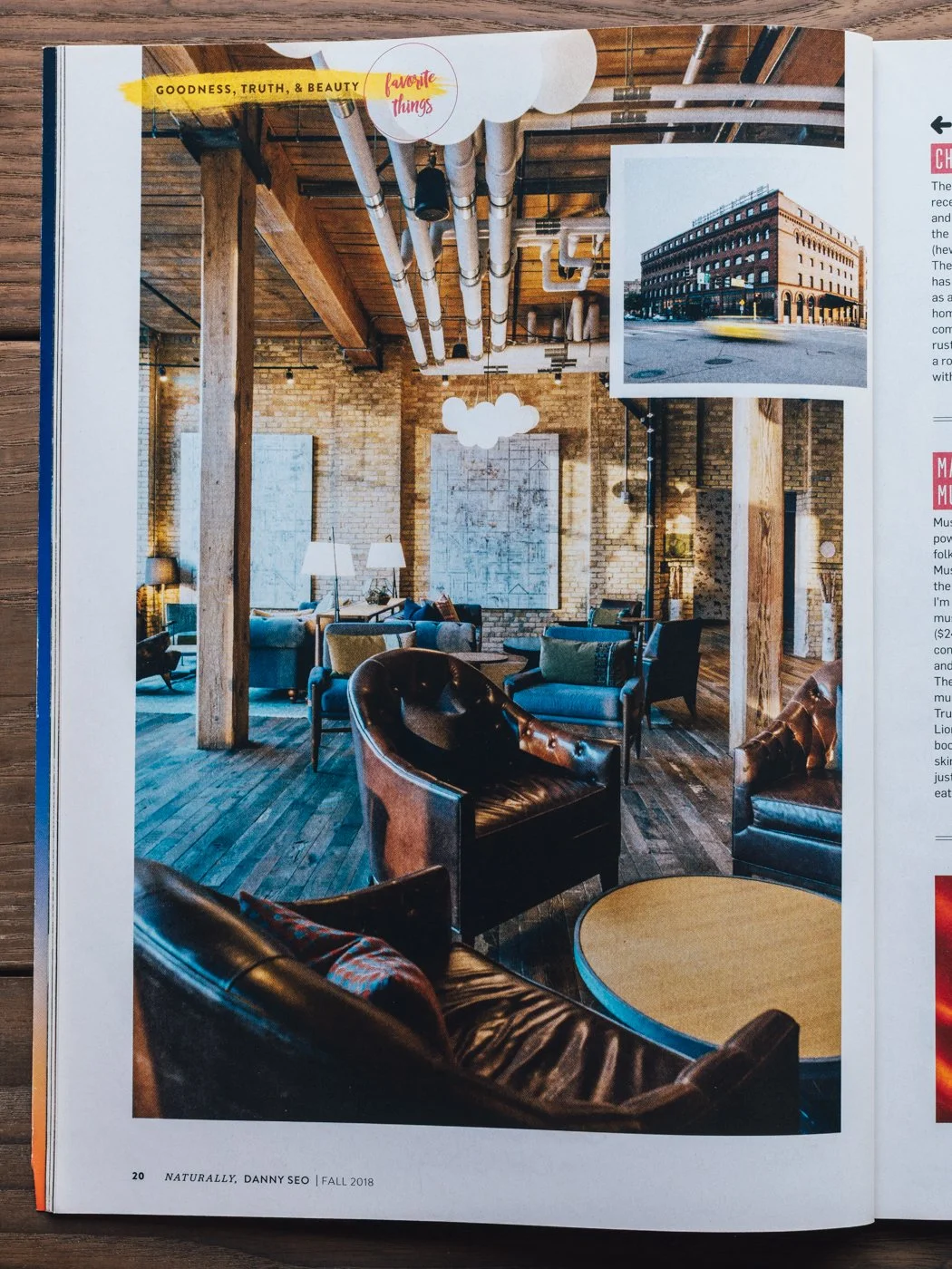 Interior view of a stylish modern lounge or cafe with exposed brick walls, wooden ceiling beams, and industrial pipes. The space is furnished with leather and upholstered chairs and sofas, with a small round table in the foreground. Decor includes fr
