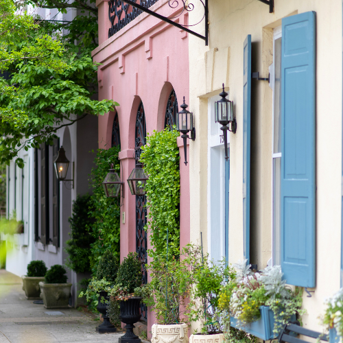 Charming pastel-colored historic homes in Charleston, South Carolina with decorative shutters, lanterns, and lush greenery along a quiet sidewalk.