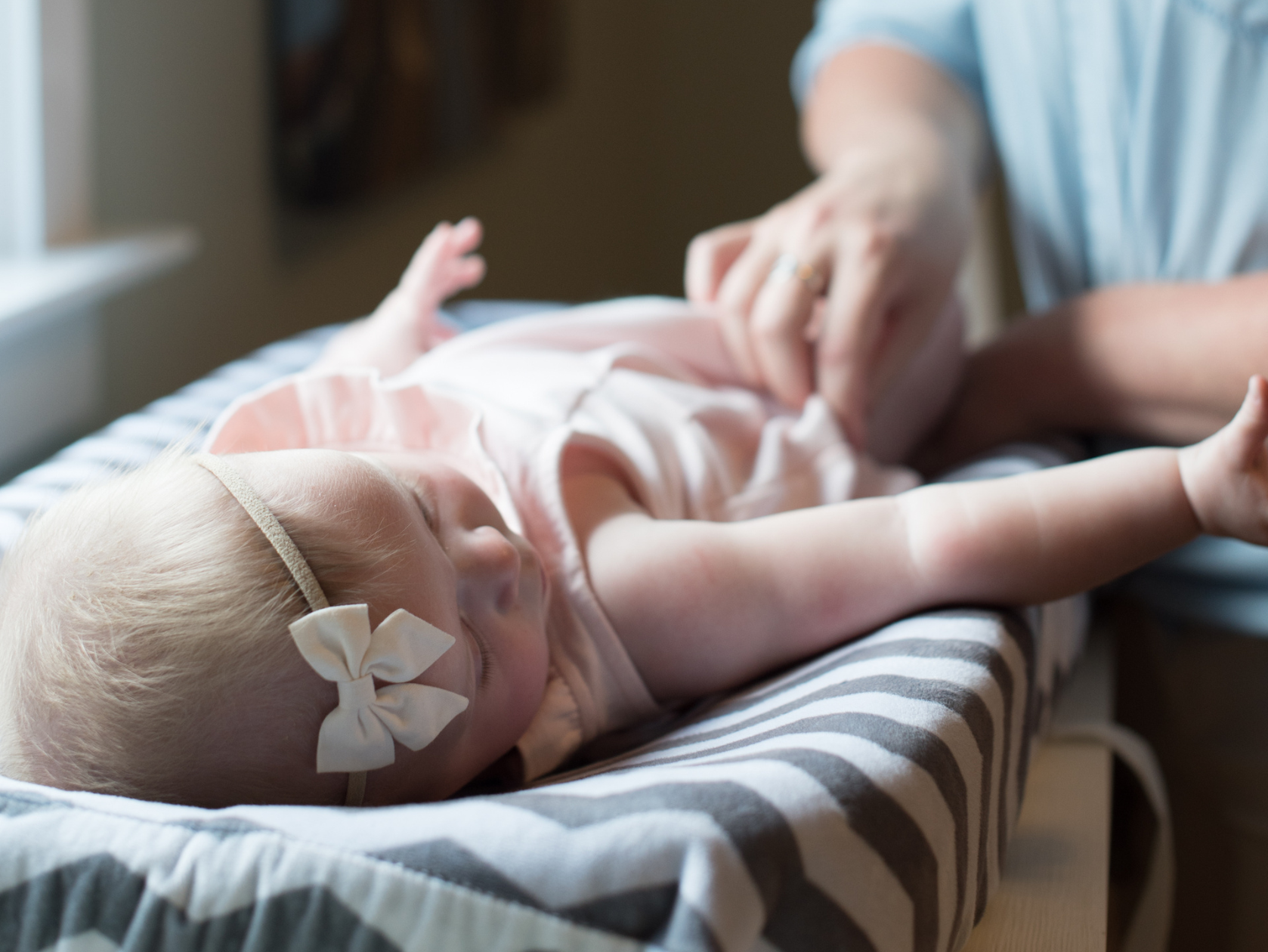 A caregiver gently changes a baby on a striped changing pad in a softly lit nursery. The infant wears a light pink outfit and a white bow headband, creating a calm and nurturing nighttime care scene.