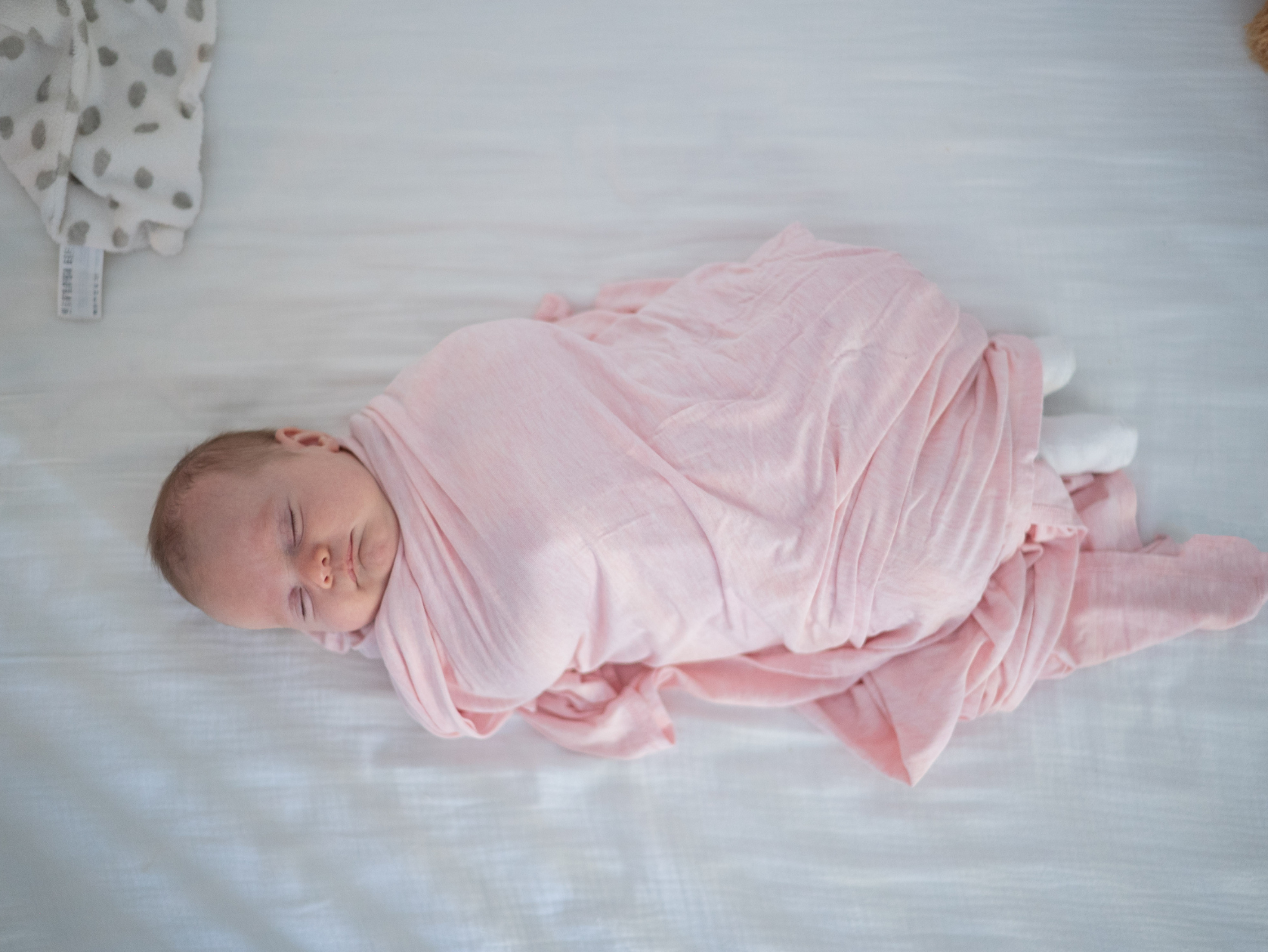 A sleeping newborn wrapped snugly in a soft pink swaddle rests peacefully on a white bed. The overhead view captures the baby’s serene expression and cozy blanket, creating a calm and comforting nursery scene.