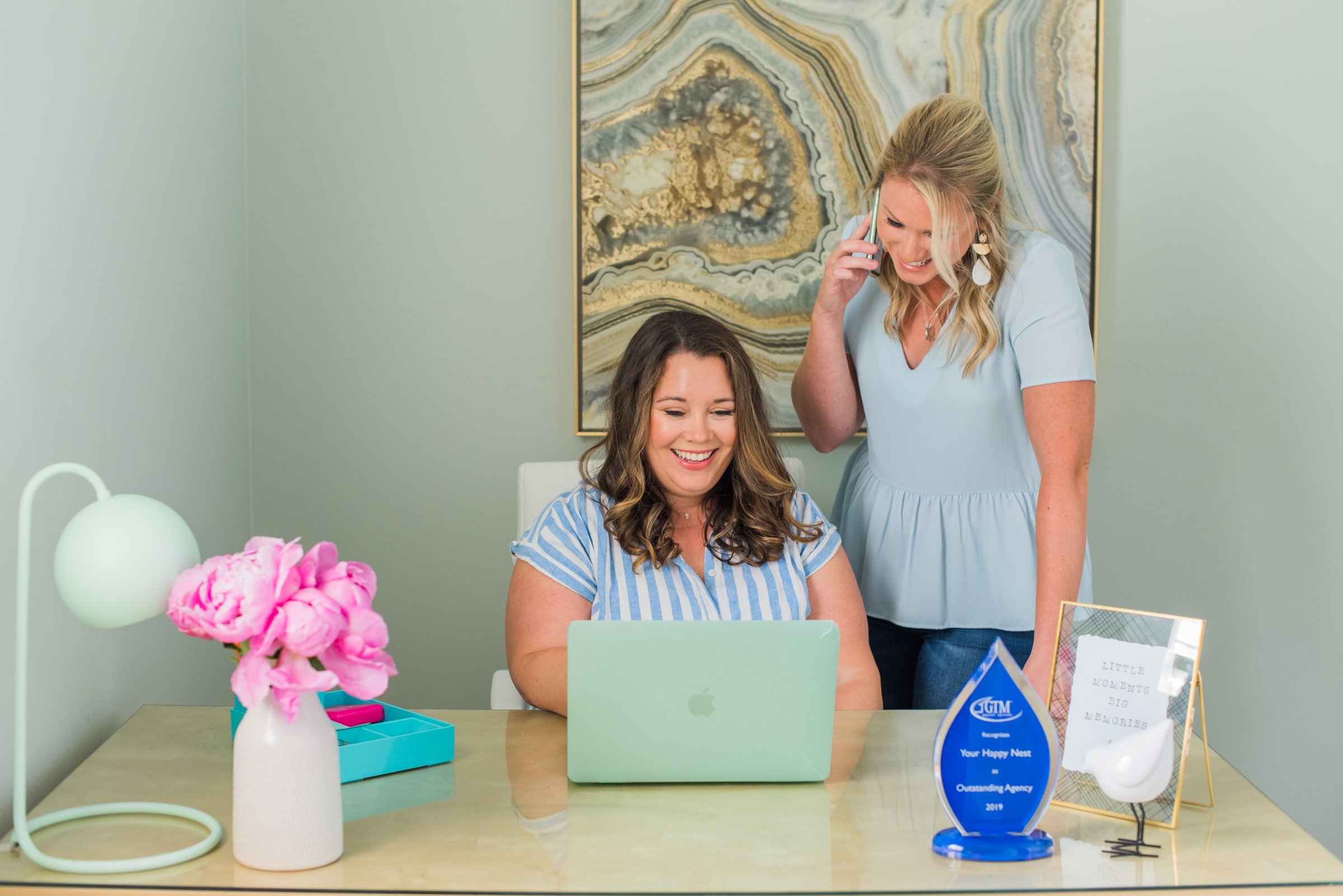 Professional childcare agency team members working together in a modern office, smiling while using a laptop and talking on the phone, with decorative artwork and flowers in the background.