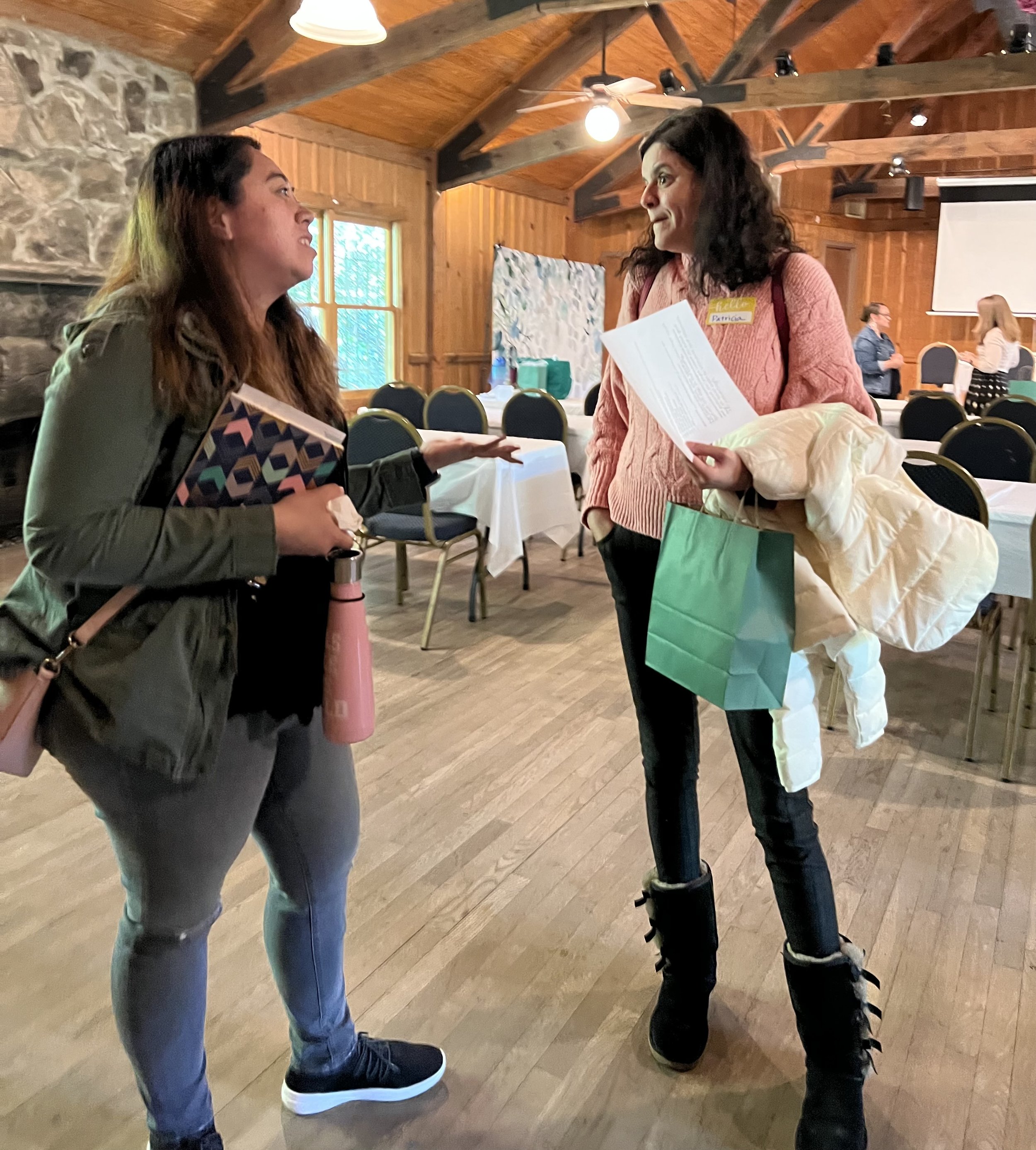 Two women conversing in a rustic wooden room with tables, chairs, and high ceilings. One woman holds a pink water bottle and a notebook, while the other carries a paper bag and a coat. There's a stone wall and window in the background.
