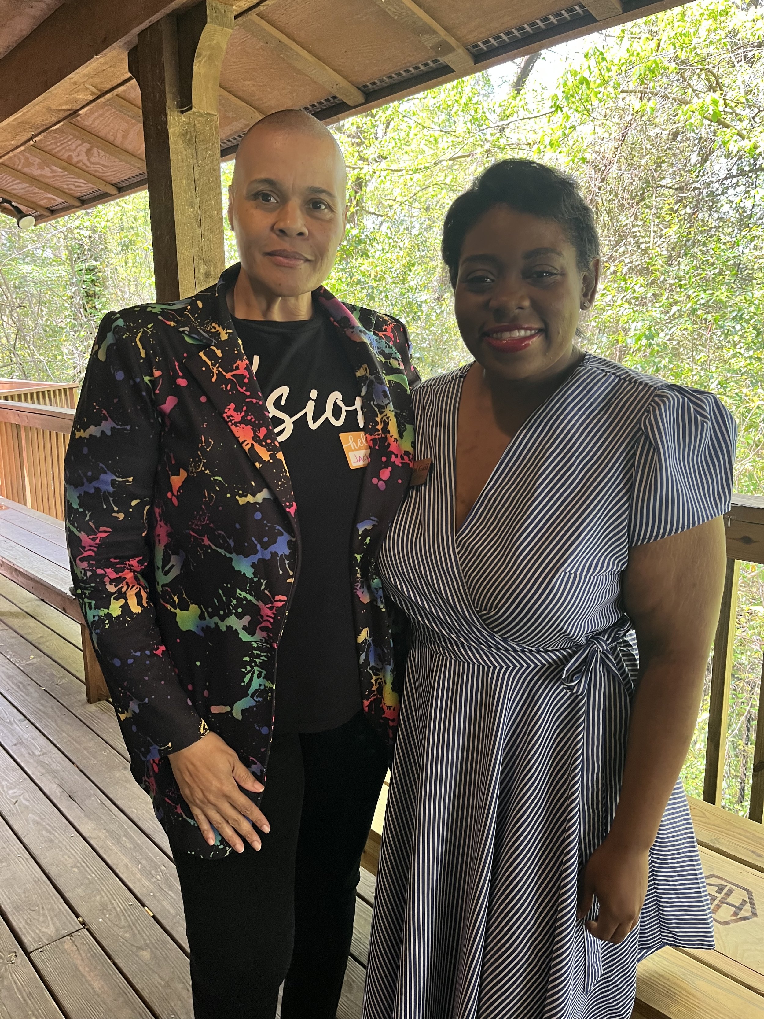 Two people standing together on a wooden deck with greenery in the background, one wearing a colorful blazer and black shirt, the other in a striped dress.
