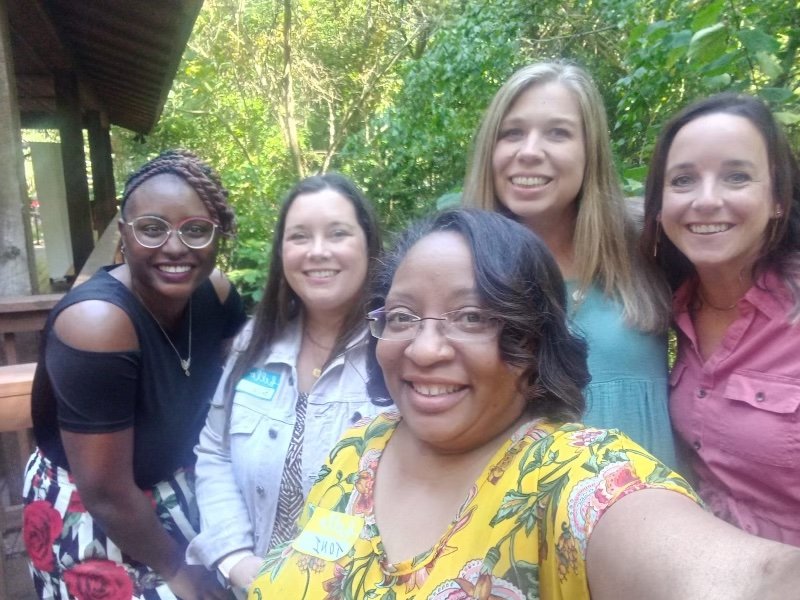 Group of five women smiling outdoors in front of trees.