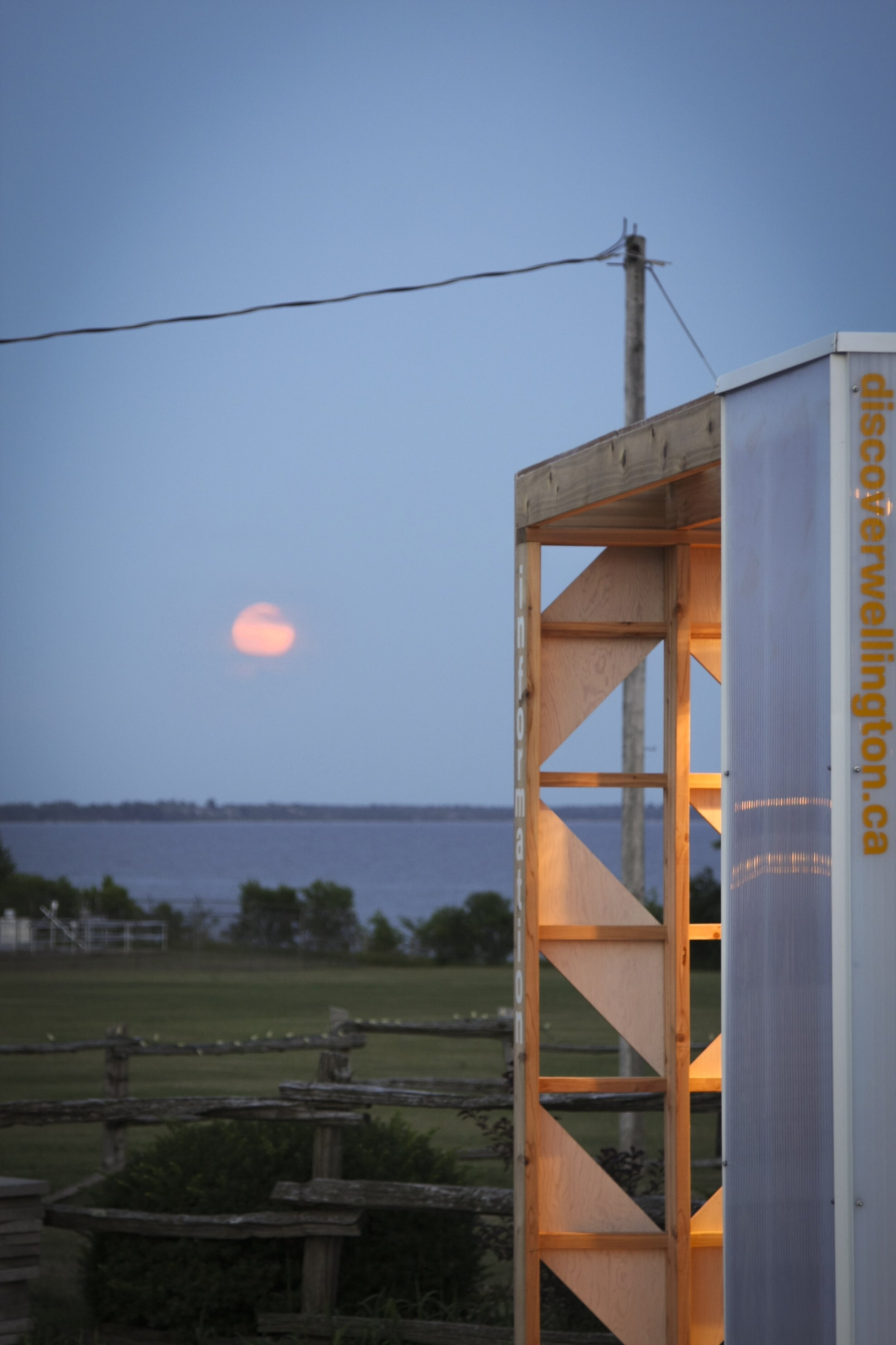 A wooden structure with a partially enclosed white side panel and yellow text, set outdoors at dusk with a view of a lake, trees, a wooden fence, and the moon in the sky.