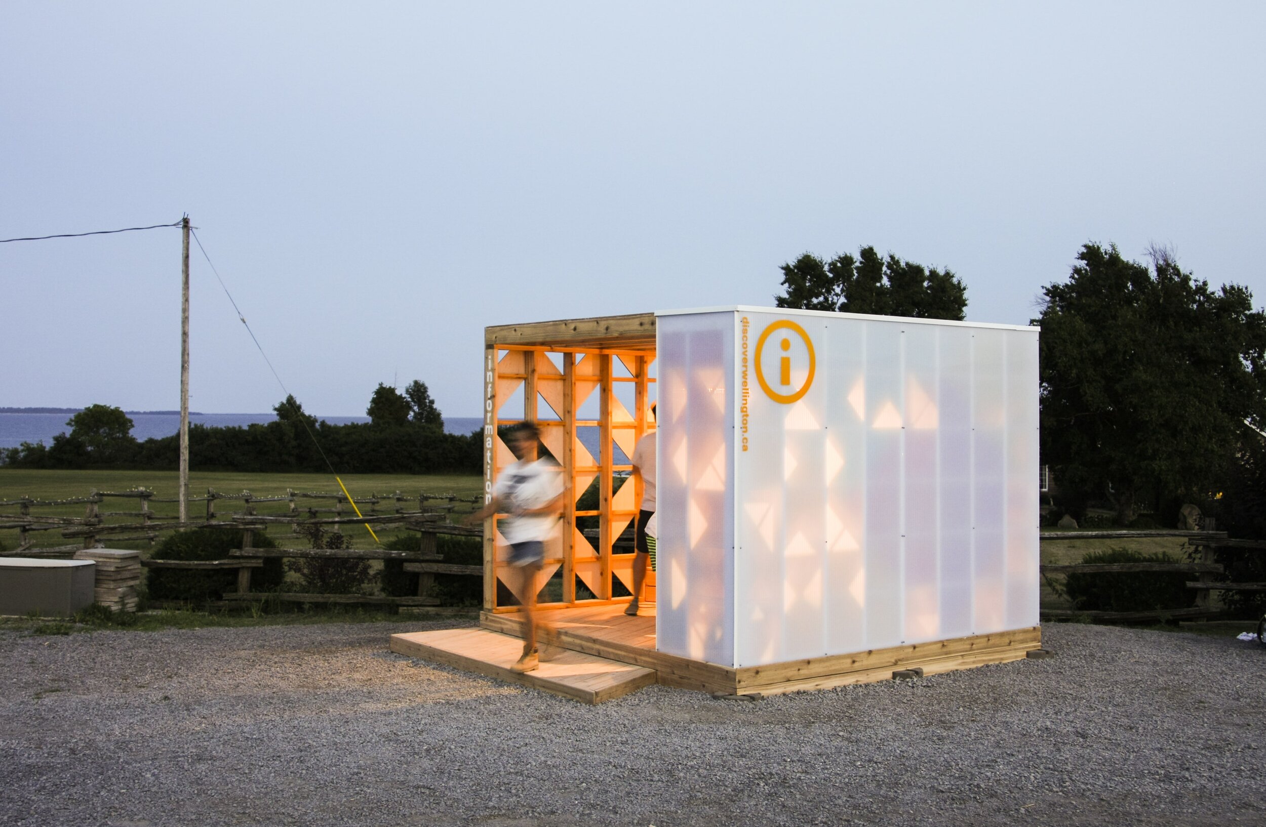 People entering a small, illuminated outdoor information booth with an orange 'i' symbol, situated on a gravel surface in a rural area with trees and open fields in the background during dusk.