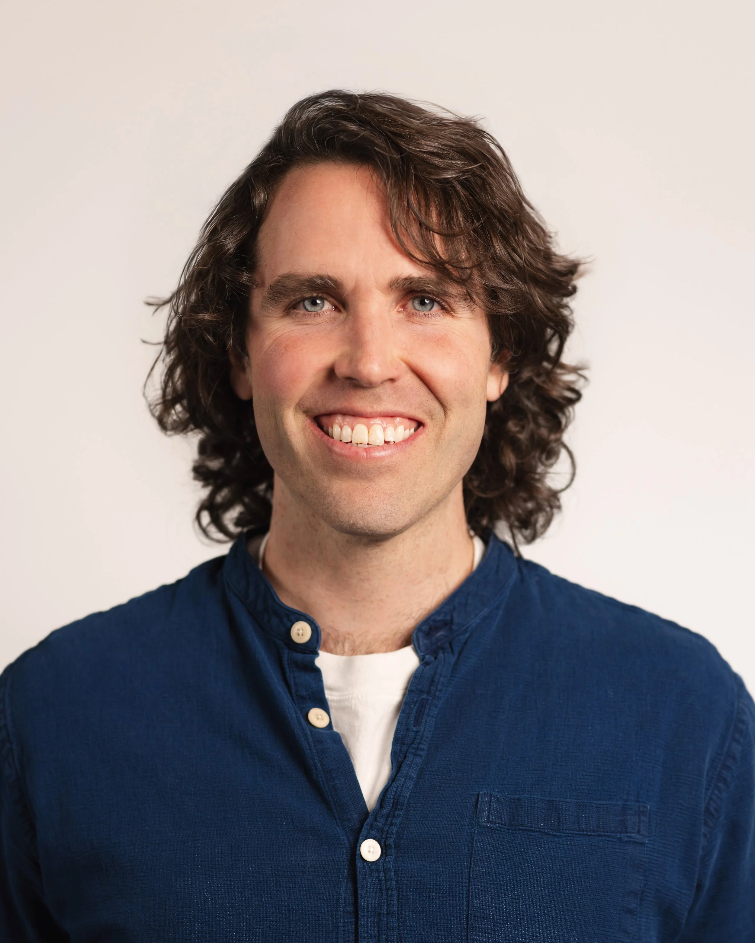 A man with long, dark, wavy hair and blue eyes, smiling, wearing a blue collared shirt and a white undershirt against a plain light background.
