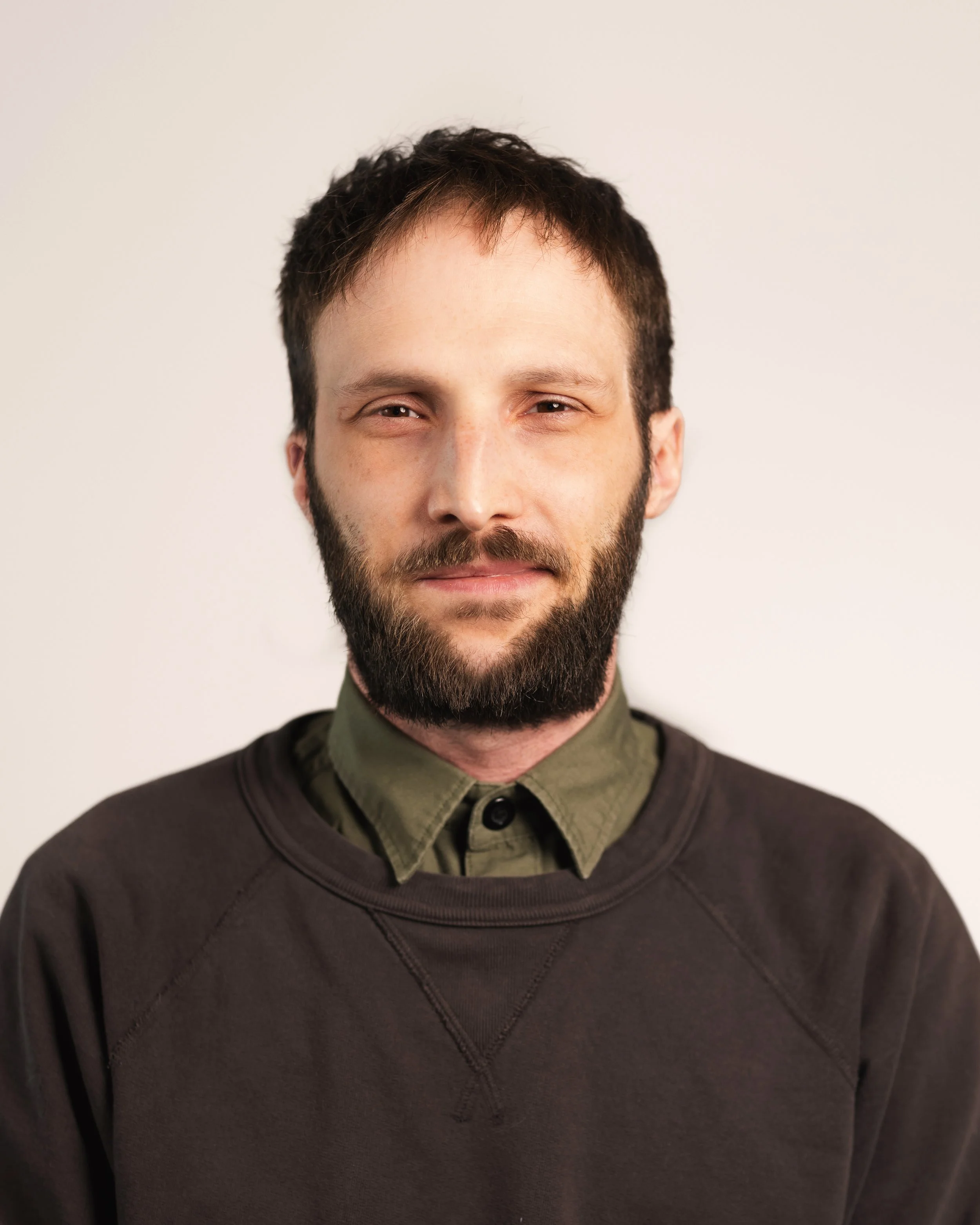 A young man with a beard and short brown hair, wearing a green collared shirt under a dark sweatshirt, smiling slightly against a plain light background.