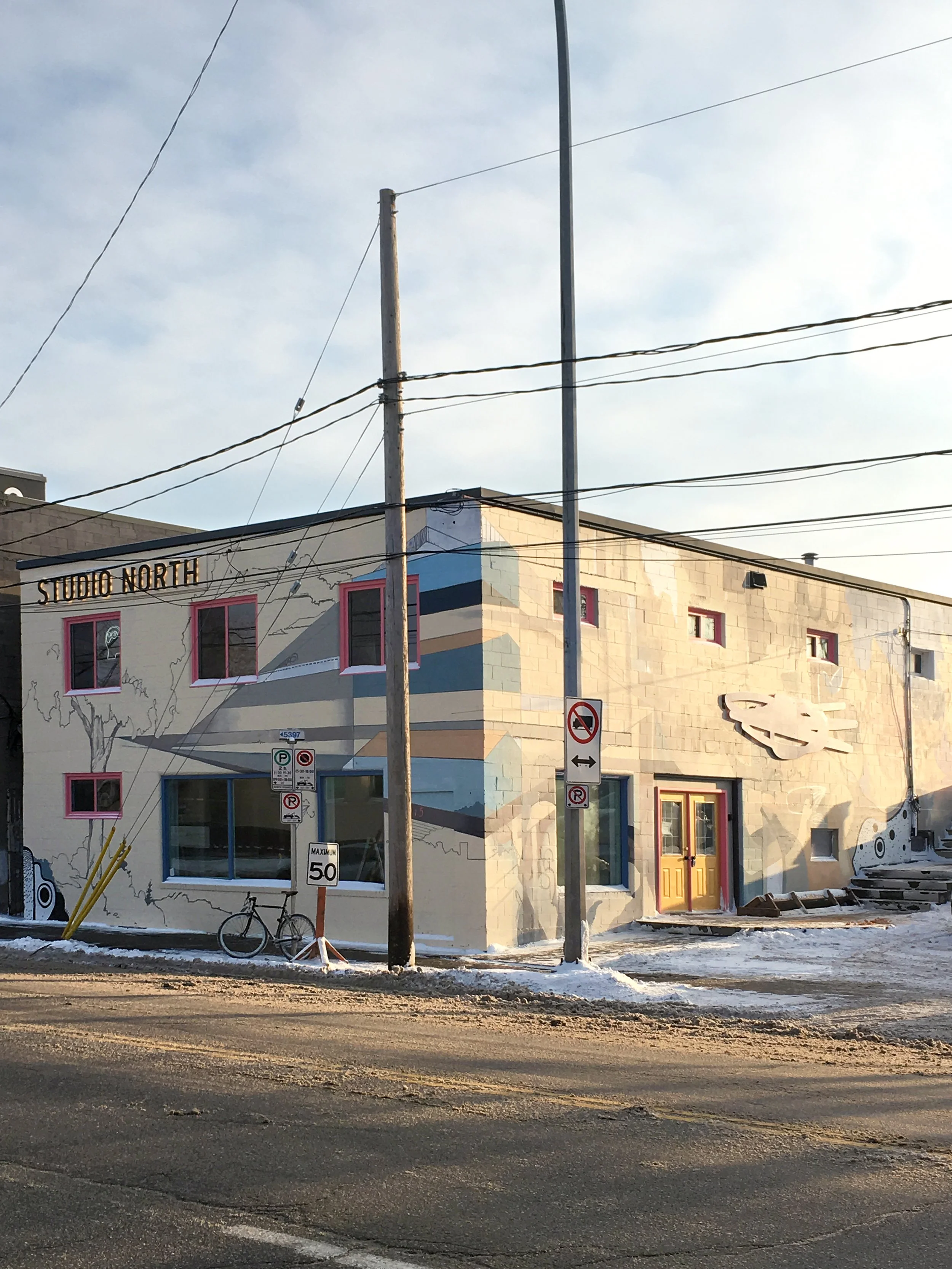 A mural on a building at Station North featuring abstract geometric shapes, a tree, a spaceship, and other designs, with the building name on top left reading "STUDIO NORTH," and street signs, a bicycle, and power lines in the foreground.