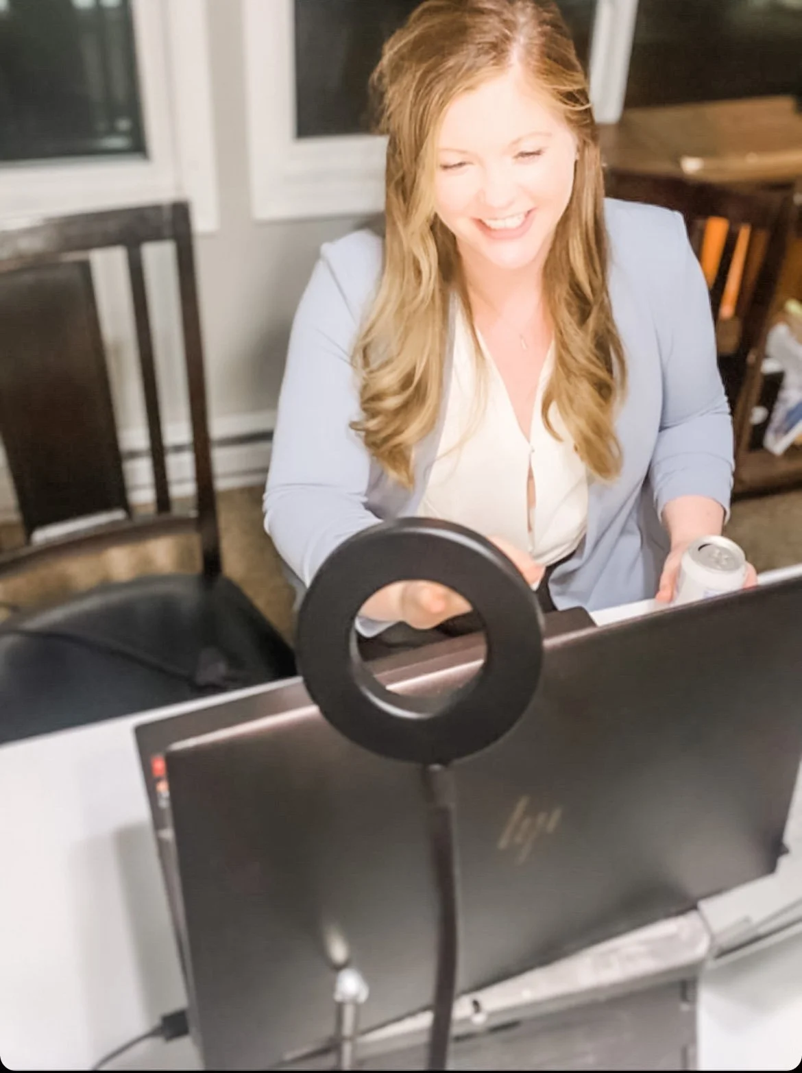 Woman smiling in front of a computer with a ring light, holding a can, in a home office setting.