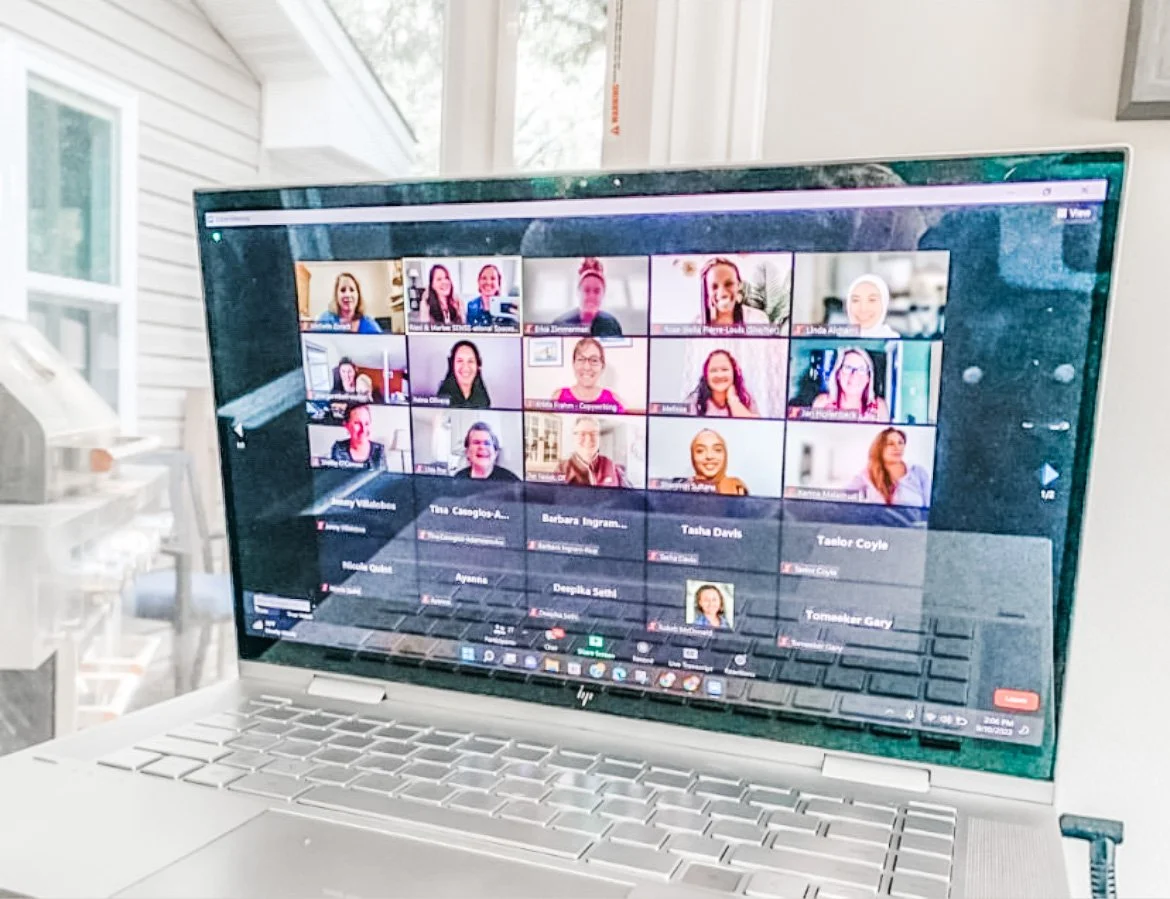 A laptop displaying a Zoom video conference with multiple participants visible on the screen. The laptop is situated on a desk next to a window with some outdoor elements visible in the background.