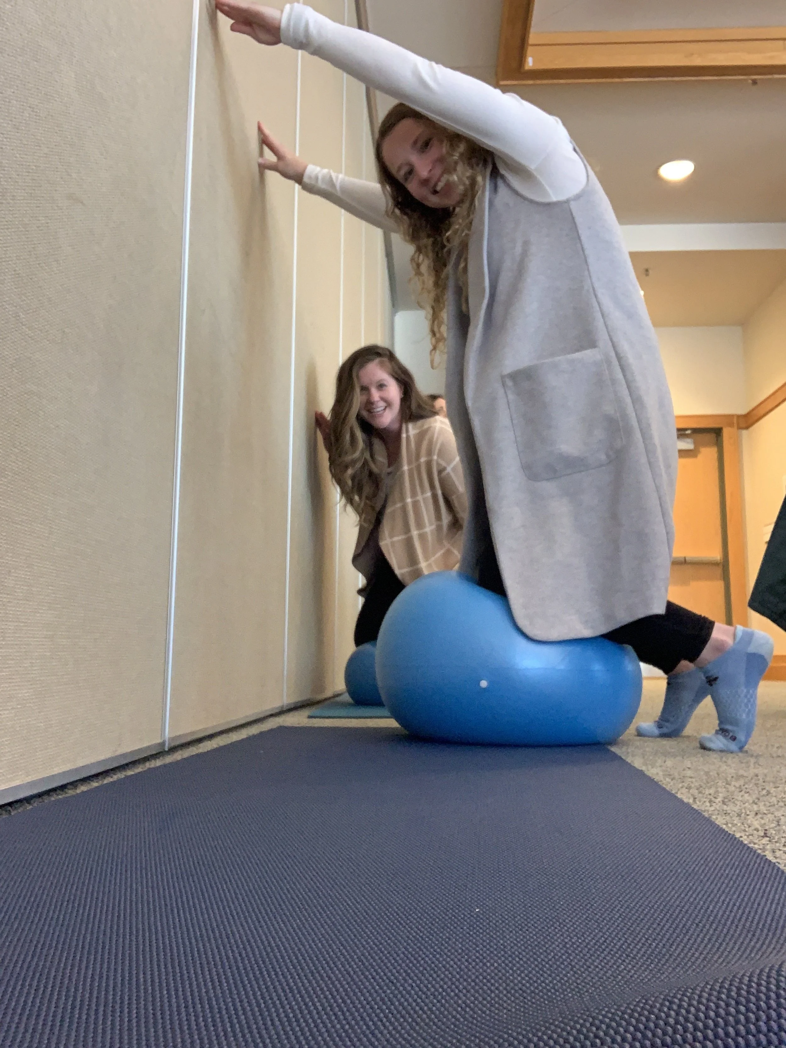 Two women using a blue exercise ball against a wall inside a room, one kneeling and the other leaning, with a yoga mat on the floor.