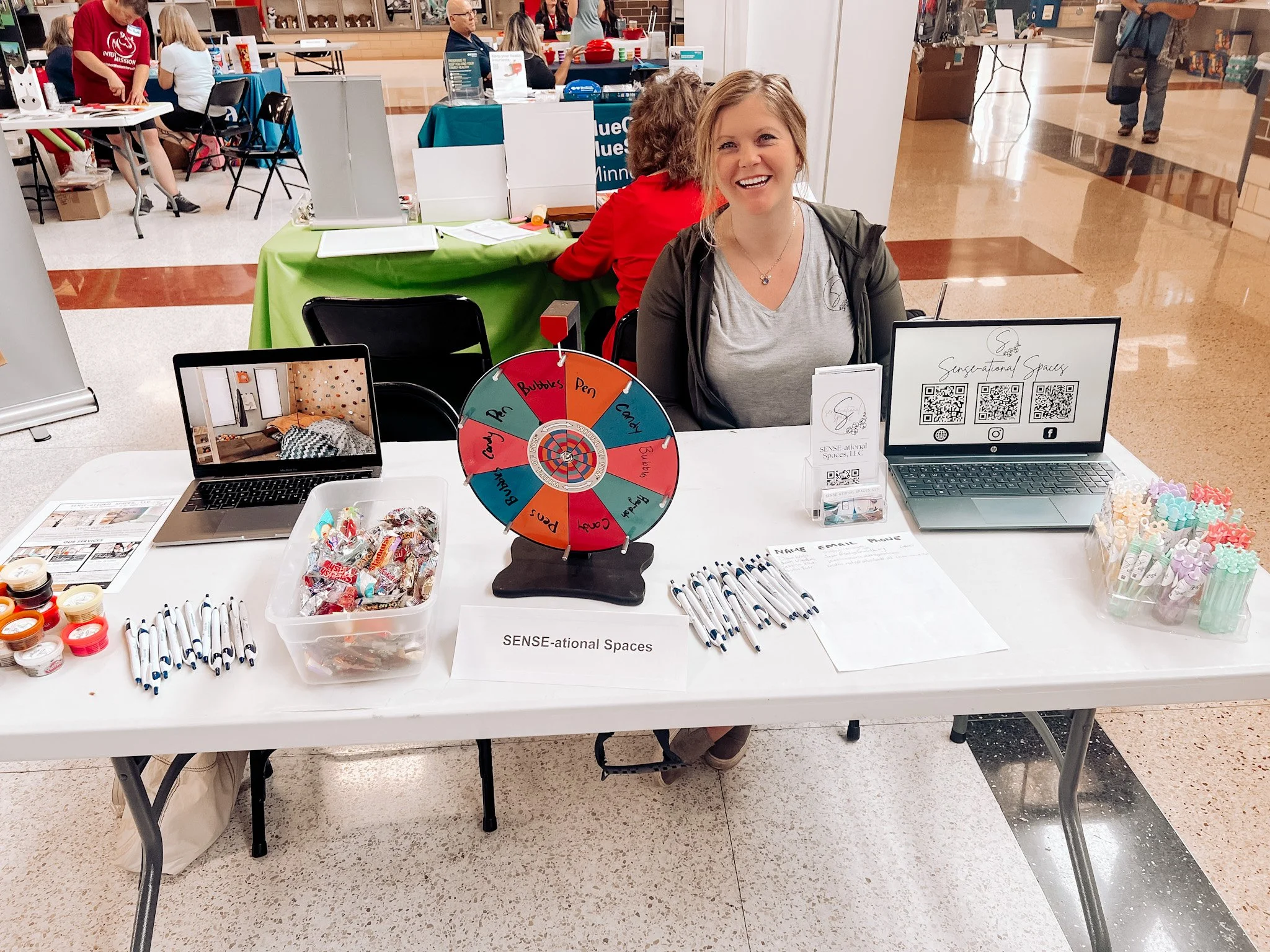A woman sits at an event booth with a display table featuring laptop, brochures, and a prize wheel labeled with options like 'Pens' and 'Bubbles.' Various items are arranged on the table, including pens, a container of candies, and small jars. The sign reads 'SENSE-ational Spaces.' Other booths and people are visible in the background.