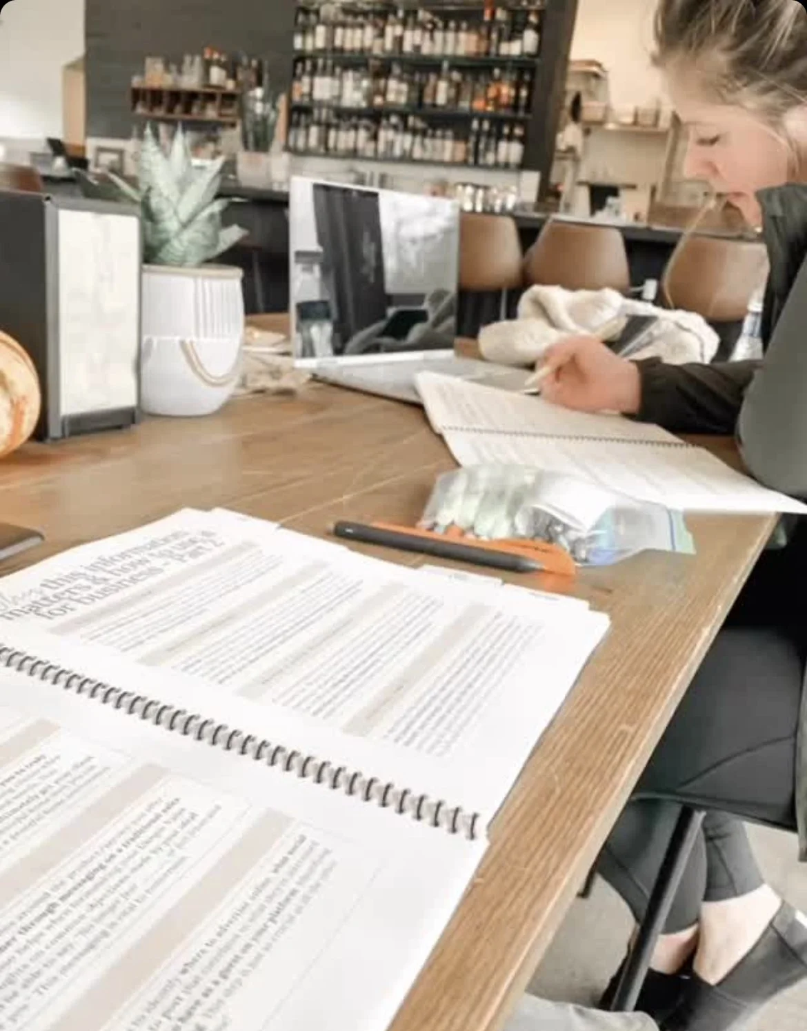 A woman studies at a café table with an open book, notebook, laptop, pen, and potted plant.