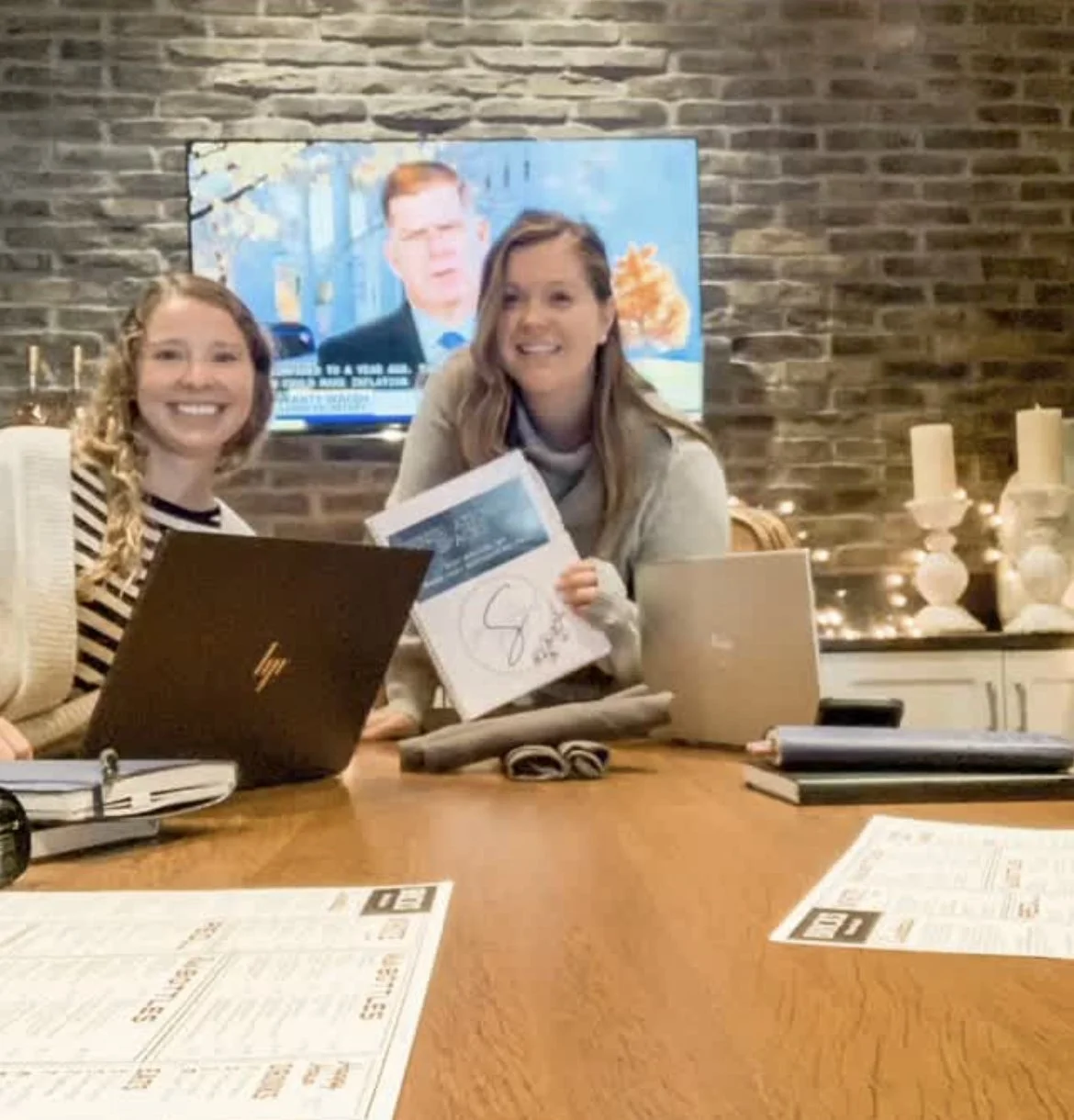 Two women sitting at a table with laptops, smiling at the camera. One woman holds a document with a signature. A TV is mounted on the brick wall behind them, showing a news broadcast. The table has notebooks, a menu, and other office items.