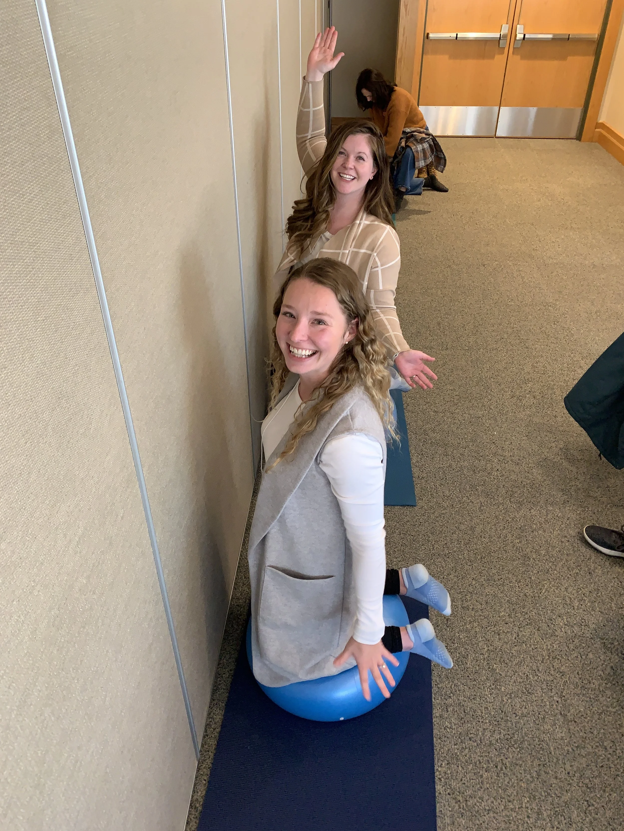 Two women sitting on exercise balls against a wall, smiling and raising their arms, with a third person in the background crouching.