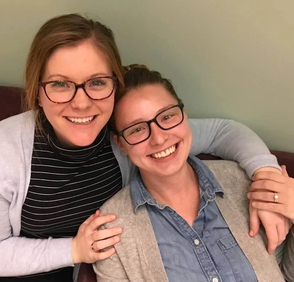 Two women wearing glasses, smiling, with one arm around each other's shoulders, seated against a light-colored background.