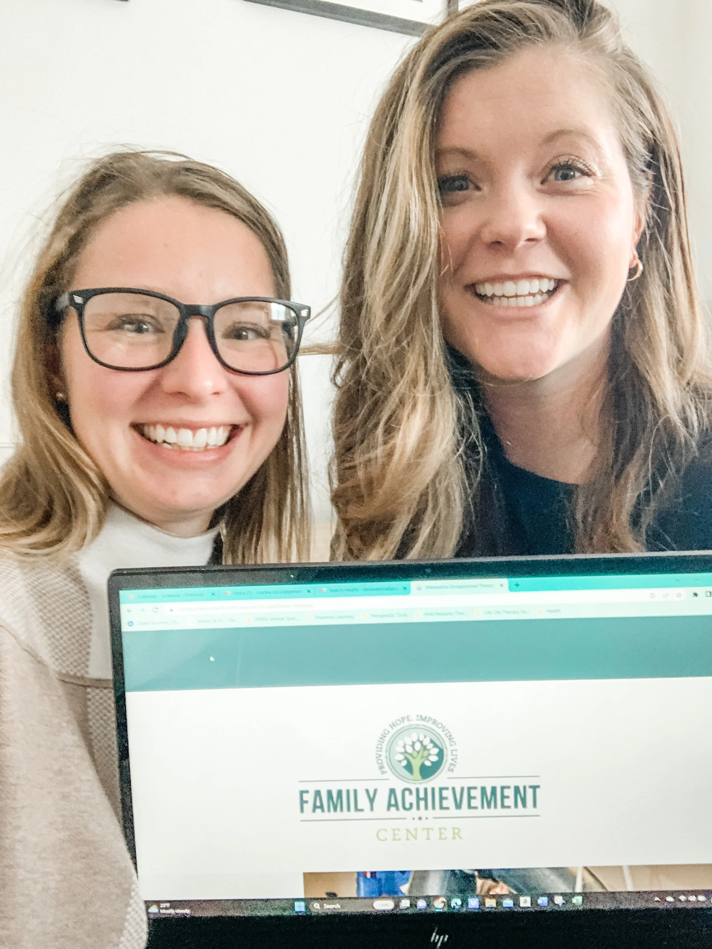 Two women smiling at the camera next to a computer screen displaying the Family Achievement Center website.