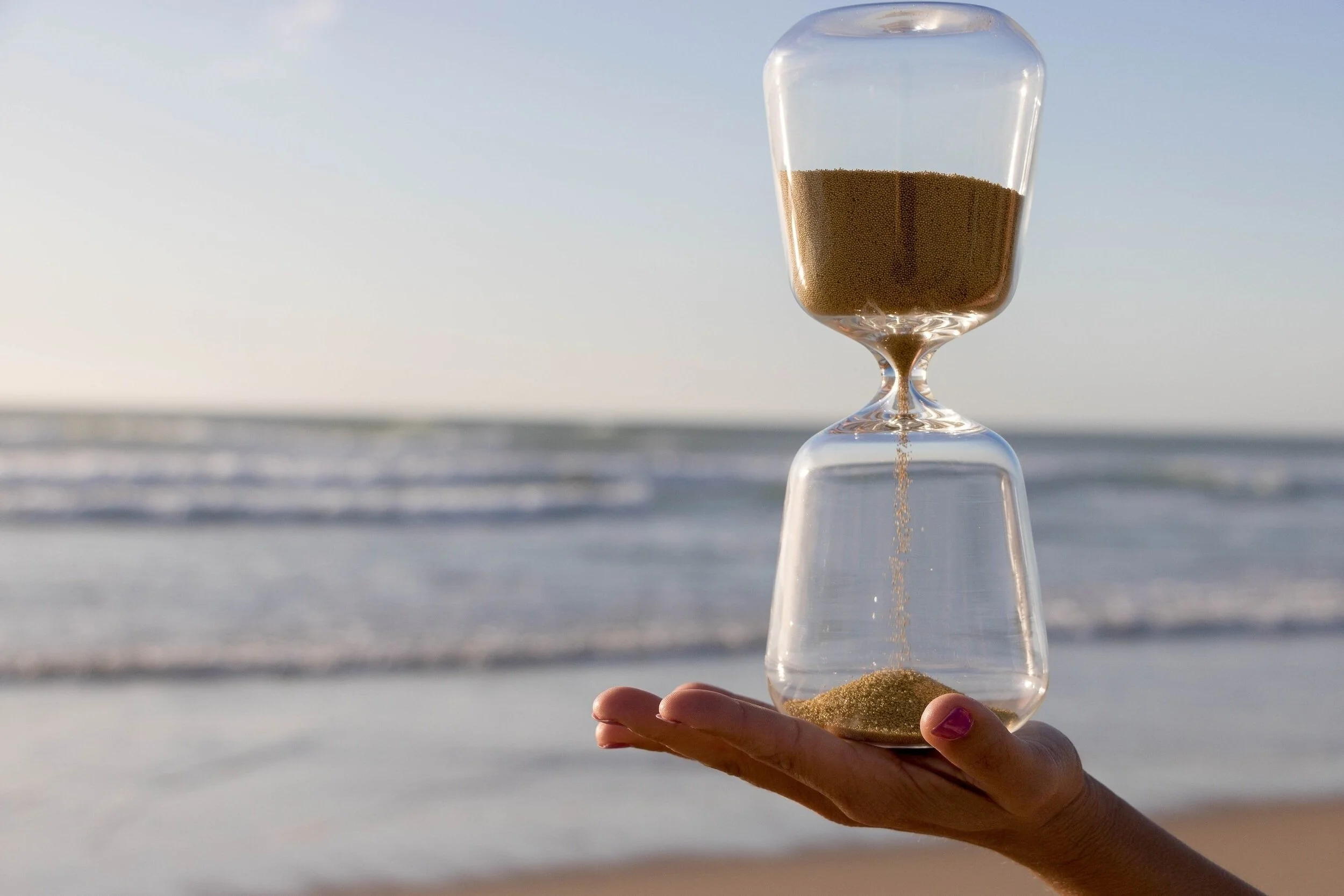 Hand holding a sand hourglass in front of a beach setting