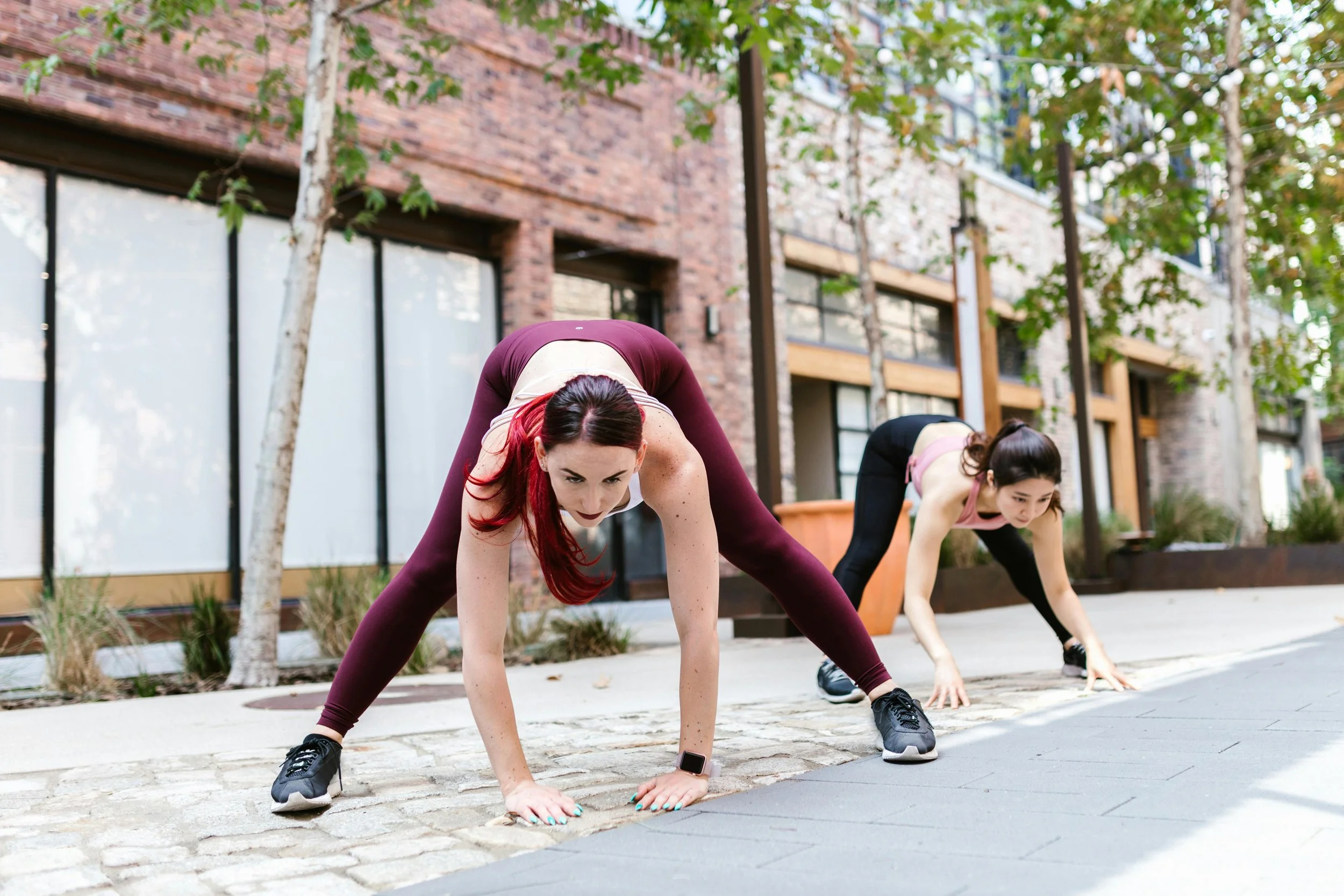 Two women in athletic wear stretch in an outdoor urban setting, surrounded by greenery and modern buildings.