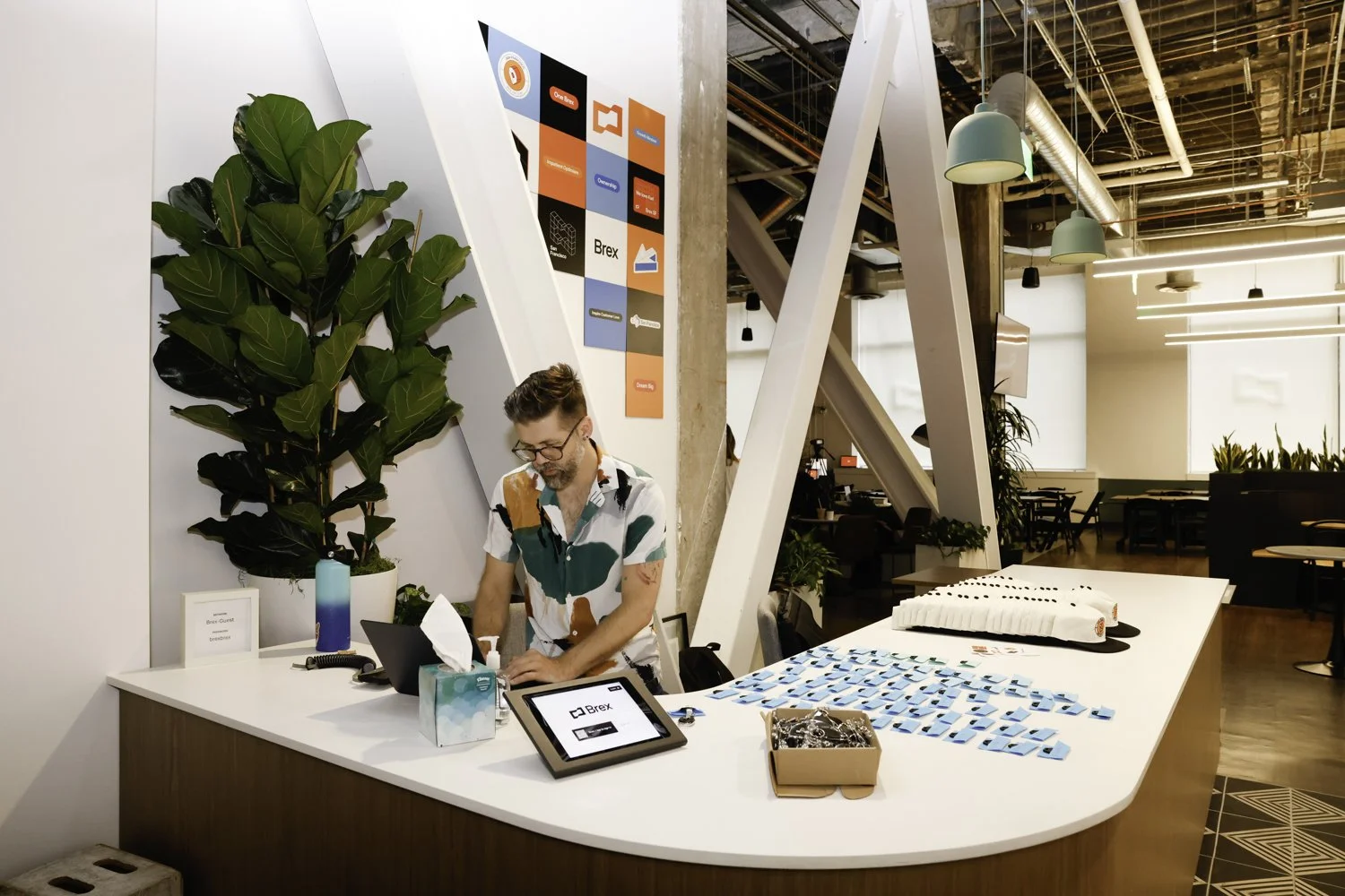 Reception desk at an office with a man working behind the counter, a large green plant, and various items including pens, a tablet, and promotional items on the counter. Modern interior with hanging lights and exposed ceiling pipes Brex headquarter i